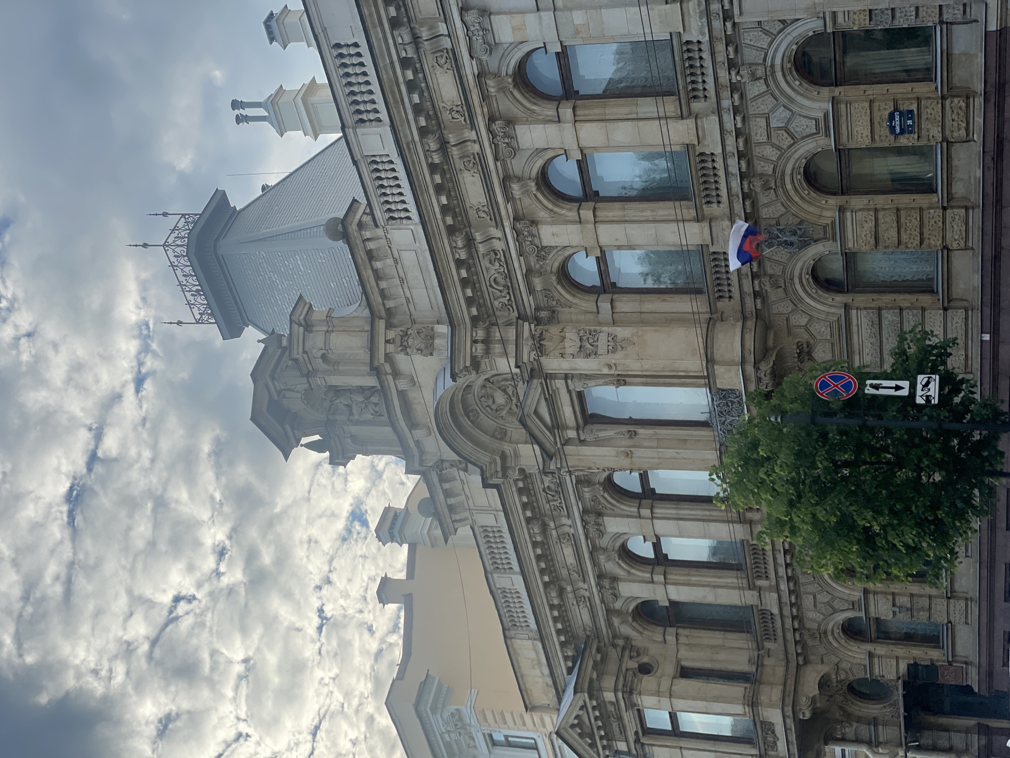 The image displays a detailed view of a grand, ornate stone building under a cloudy sky, rotated 90 degrees clockwise. The architecture features a heavily decorated facade with intricate carvings, decorative friezes, and numerous arched windows with reflective glass. Above the main structure, a mansard roof with elaborate metalwork cresting and chimneys is visible against the overcast sky.
No people are present in the scene, which appears static. The time of day is daytime, indicated by the overall brightness despite the dense, white and gray clouds.
Notable details include a small Russian flag (white, blue, and red horizontal stripes) hanging from the building's facade. Directly below the flag, a blue street sign with white Cyrillic text is clearly visible, reading "Чайковского" (Chaykovskogo) and the number "28", indicating the street name and building number. A lush green tree partially obscures the lower part of the building, and a red circular street sign (likely a "no stopping" sign) along with an arrow sign are partially visible near the tree. The architectural style, the Russian flag, and the Cyrillic street sign firmly place the location in Saint Petersburg, Russia, aligning with the provided context.