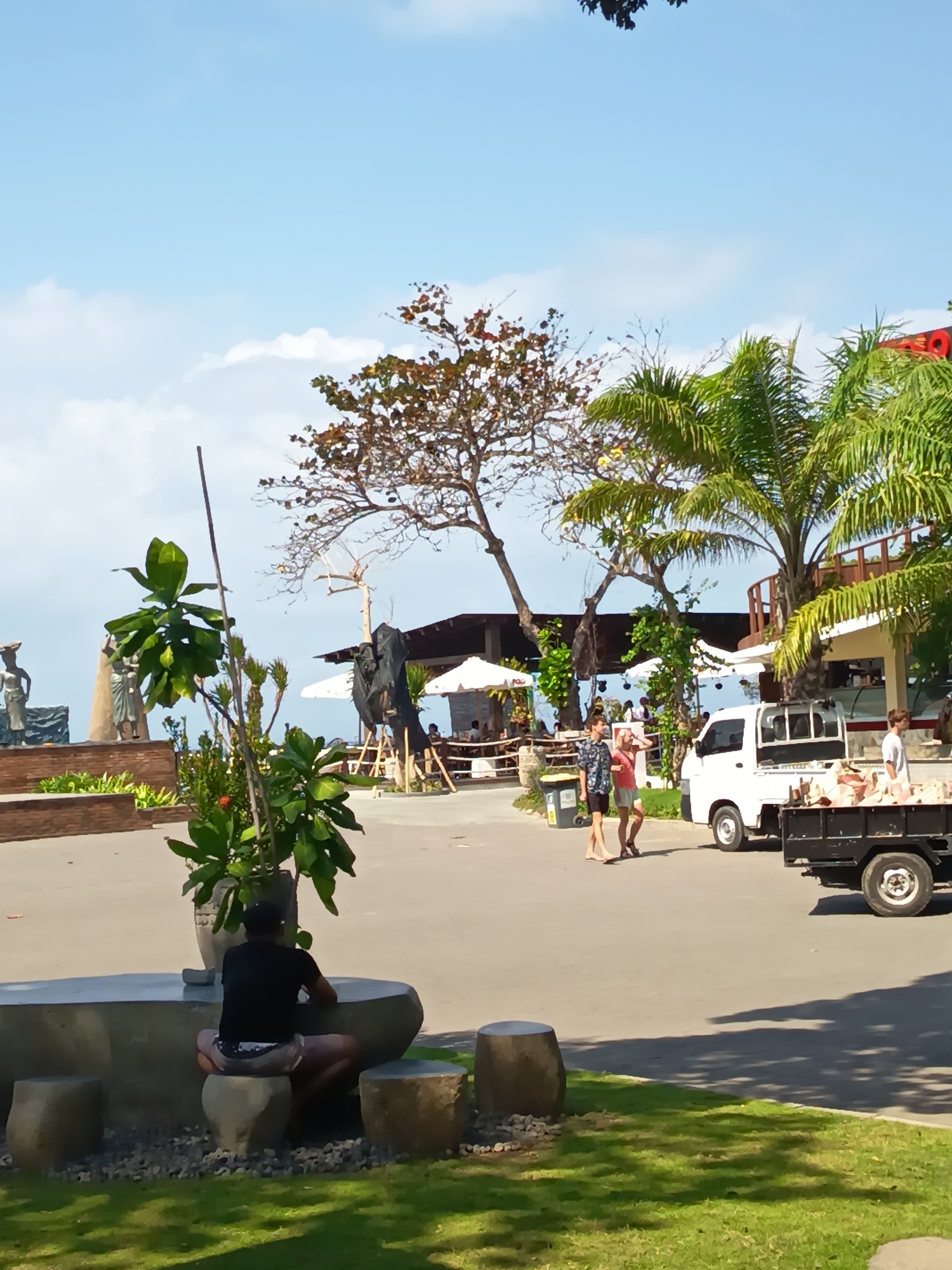 The image captures a scene in Denpasar, Indonesia, under a clear blue sky with scattered white clouds. In the foreground, a person with dark hair and a black t-shirt sits on a round, stone stool, facing away from the camera. They are positioned in front of a long, curved stone bench, with other round stools visible to the sides. Lush green grass covers the lower portion of the frame, with dappled sunlight casting shadows.
To the left, a large potted plant with broad green leaves stands near the stone bench. Further back and to the left, several statues are visible, appearing to be part of a decorative display. Behind the statues and potted plant, a large, bare-branched tree with some reddish-brown leaves stands prominently.
In the mid-ground, a white pickup truck is parked on an asphalt surface. The truck bed is filled with what appears to be light-colored goods, possibly food items or building materials. Two people, a man and a woman, are walking on the asphalt in front of the truck. The man wears a patterned shirt and dark shorts, while the woman is in a pink top and light-colored shorts.
Further into the background, there are several outdoor seating areas with umbrellas and awnings, suggesting a restaurant or cafe. Palm trees with vibrant green fronds are visible, adding to the tropical ambiance. The overall scene depicts a lively outdoor environment, likely a public space or a commercial establishment near the coast, given the presence of palm trees and the distant water visible through some openings. The sun appears to be high in the sky, indicating it is daytime.