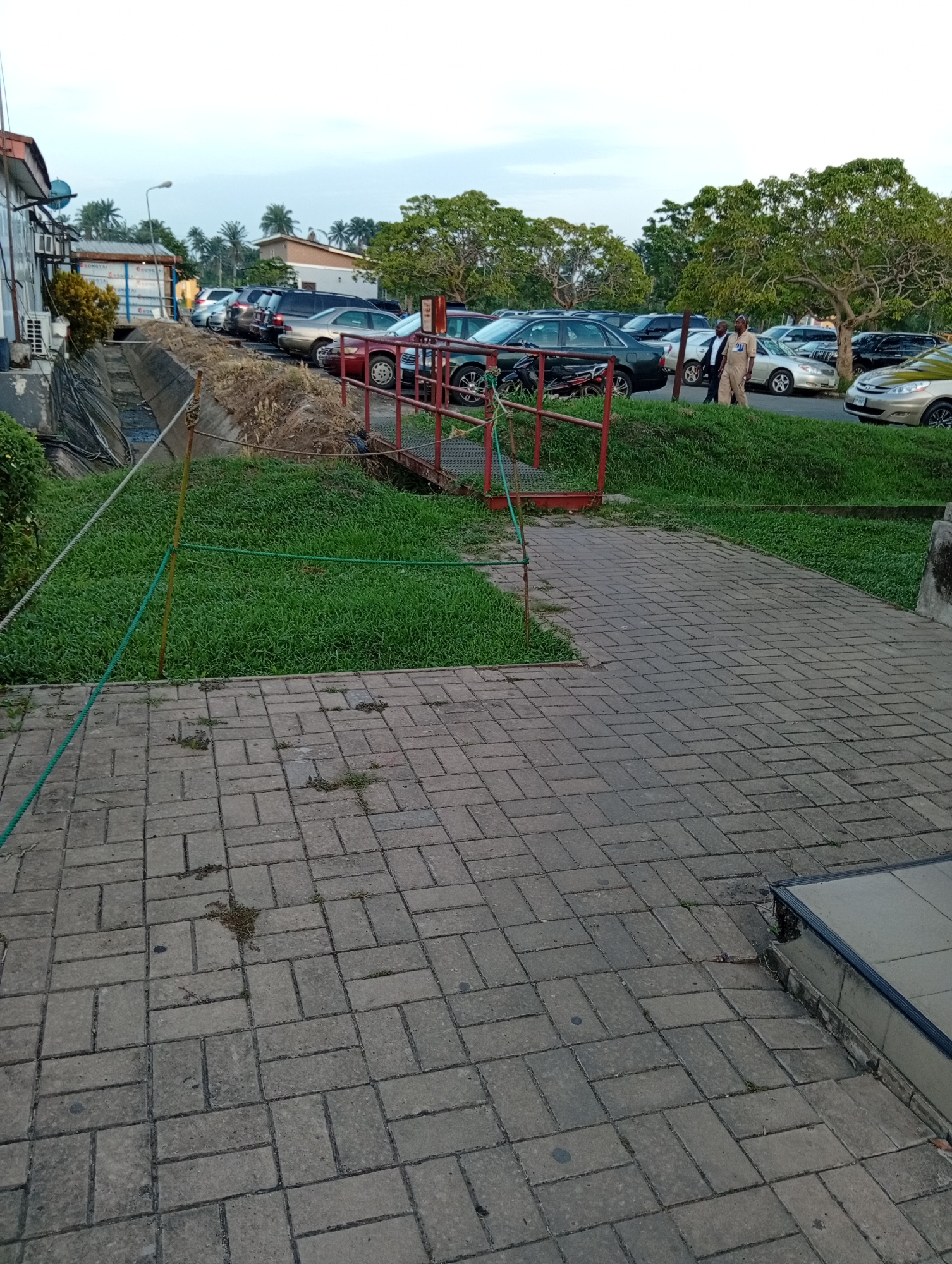 The image depicts an outdoor scene in what appears to be a parking lot or public area in Omagwa, Nigeria. The foreground is a paved walkway made of rectangular blocks. To the left, a narrow irrigation ditch separates the walkway from a building. A small, red metal bridge spans the ditch. Green grass covers the banks of the ditch, and a rope barrier runs along the edge of the walkway. In the background, several parked cars are visible, and two men in light-colored uniforms or suits are walking along the edge of the parking lot. Trees are scattered throughout the scene, providing some shade. The sky is overcast.