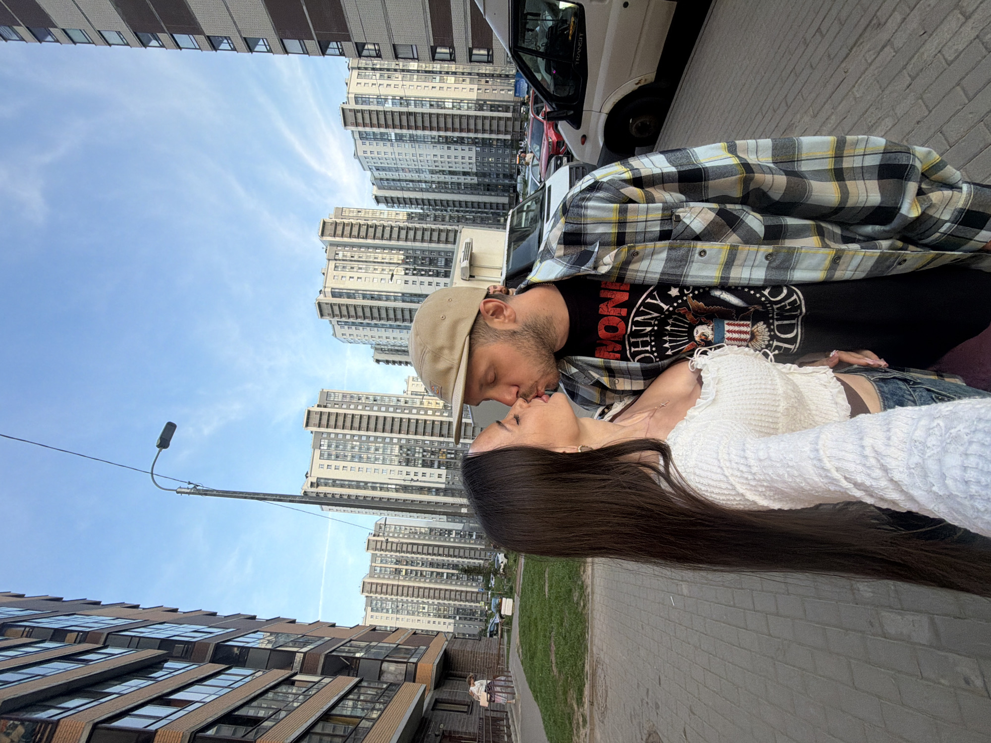 A young couple is shown kissing on a paved sidewalk in front of a row of tall, modern apartment buildings in Saint Petersburg, Russia. The woman, on the left, has long, dark brown hair and is wearing a white, off-the-shoulder top. The man, on the right, is wearing a tan baseball cap, a black t-shirt with a graphic print, and a plaid shirt. In the background, several other apartment buildings are visible, along with parked cars and a white van. The sky is blue with some wispy clouds. A streetlamp is visible to the left of the couple.