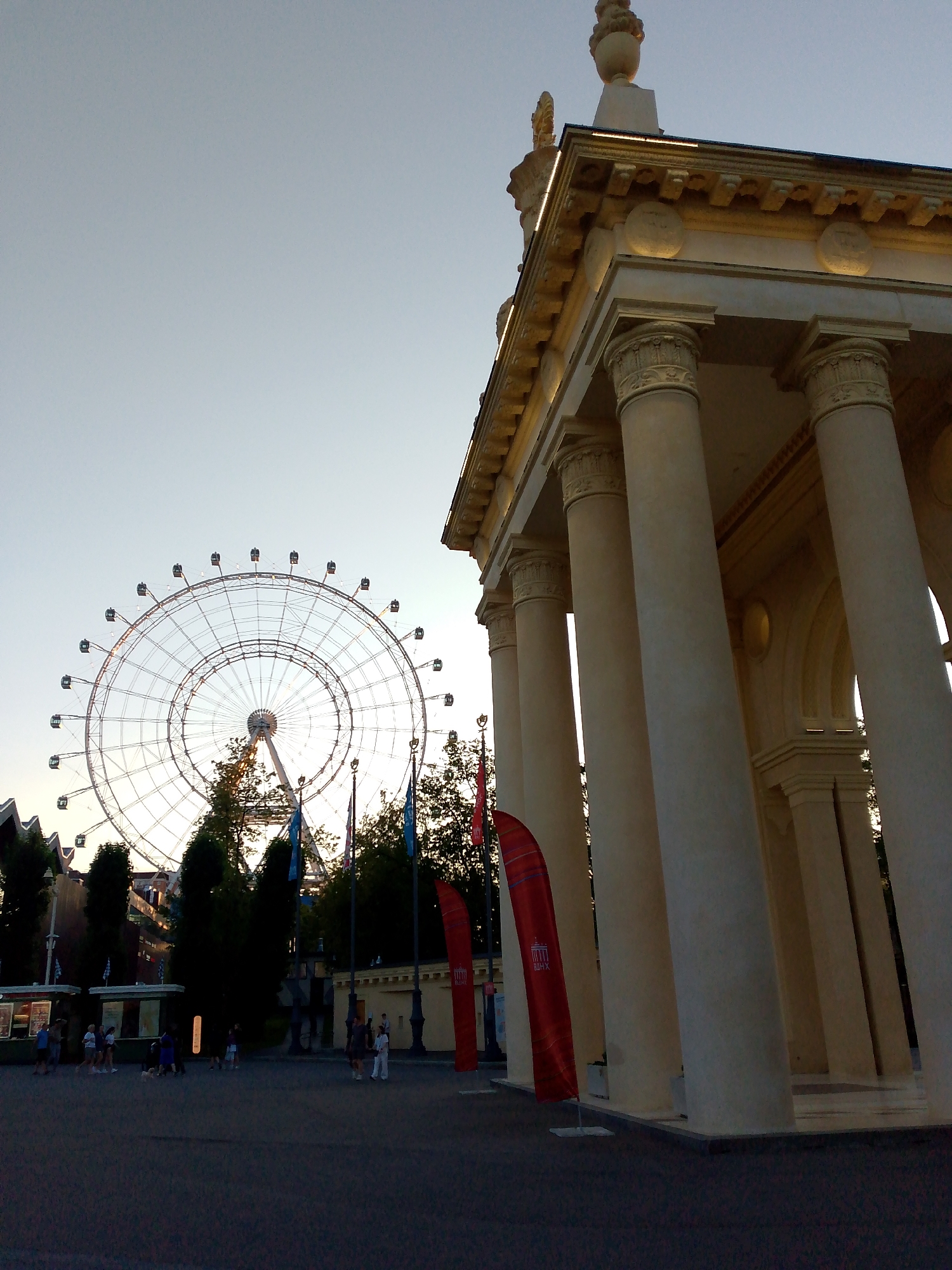 The image captures a wide-angle view of an amusement park in Moscow, Russia, likely VDNH (Exhibition of Achievements of National Economy). On the right, a grand classical pavilion with large columns and ornate detailing is prominently featured, bathed in the warm light of what appears to be late afternoon or early evening. To the left, a large Ferris wheel stands tall against the clear sky, its structure illuminated and surrounded by trees. The foreground shows a paved area where several people are walking and standing, some near food stalls or information booths. Red flags with a stylized architectural emblem are planted in the ground, adding color to the scene. The overall atmosphere suggests a public recreational space with a blend of historical architecture and modern entertainment.