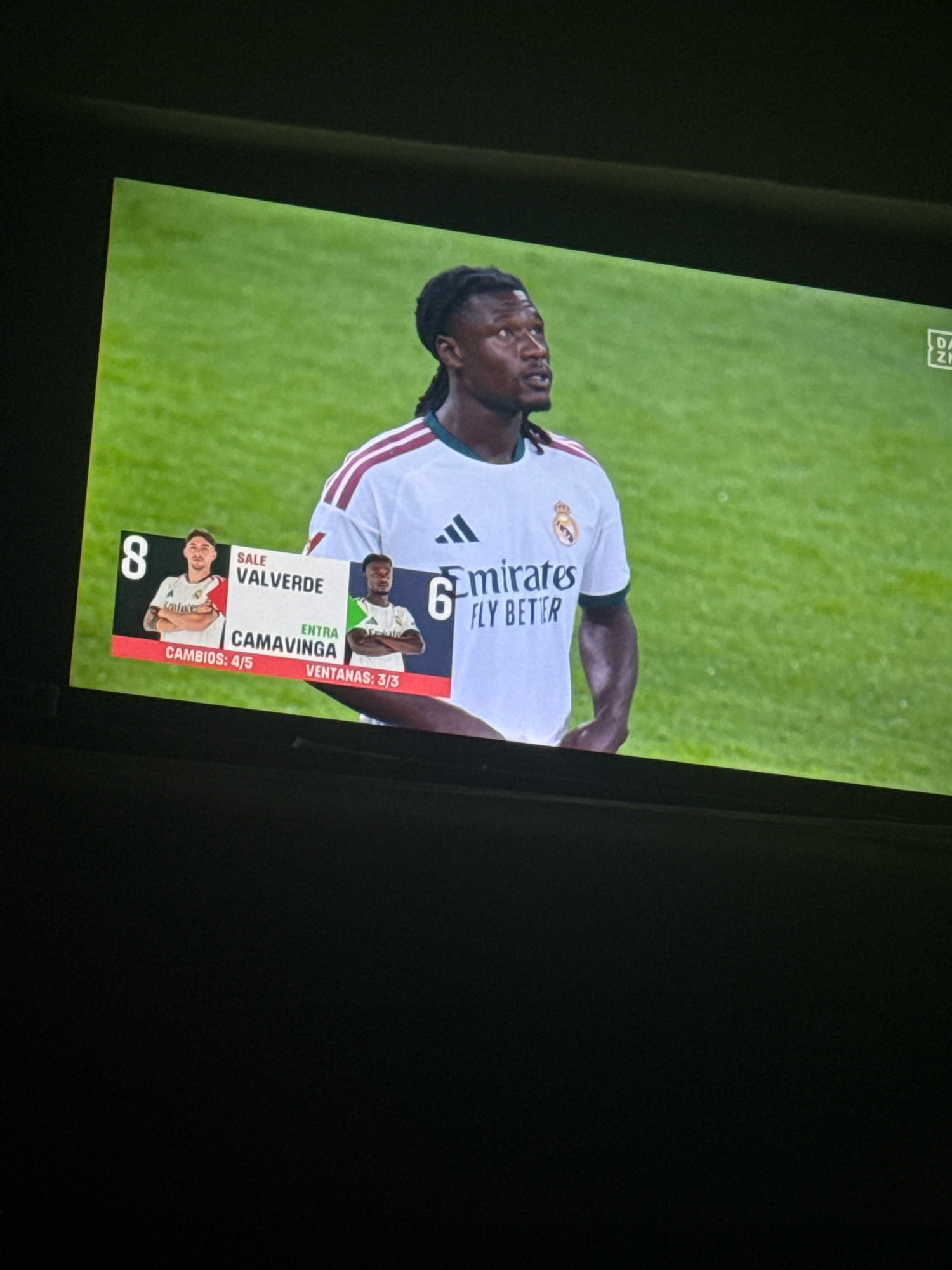 The image is a photograph taken of a television screen displaying a football match. The screen shows a close-up of a dark-skinned male football player wearing a white jersey with red and green trim. He has a serious expression and his hair is styled in dreadlocks. Below the player, there is a graphic overlay displaying information about player substitutions. On the left side of the graphic, the number '8' is visible, with a picture of another player and the name "VALVERDE" above it. To the right of this, the word "ENTRA" (entering) is written above "CAMAVINGA". Further to the right, the number '6' is visible, and the phrase "FLY BETTER" is partially visible on the player's jersey. The background of the television screen is a green football pitch. The photograph is taken at an angle, and the bottom portion of the image is black, suggesting the screen is framed by a television set. The overall scene suggests a live sports broadcast. The specific location of Chañe, Spain, is not depicted in the image itself, but is provided as context for the event being broadcast.