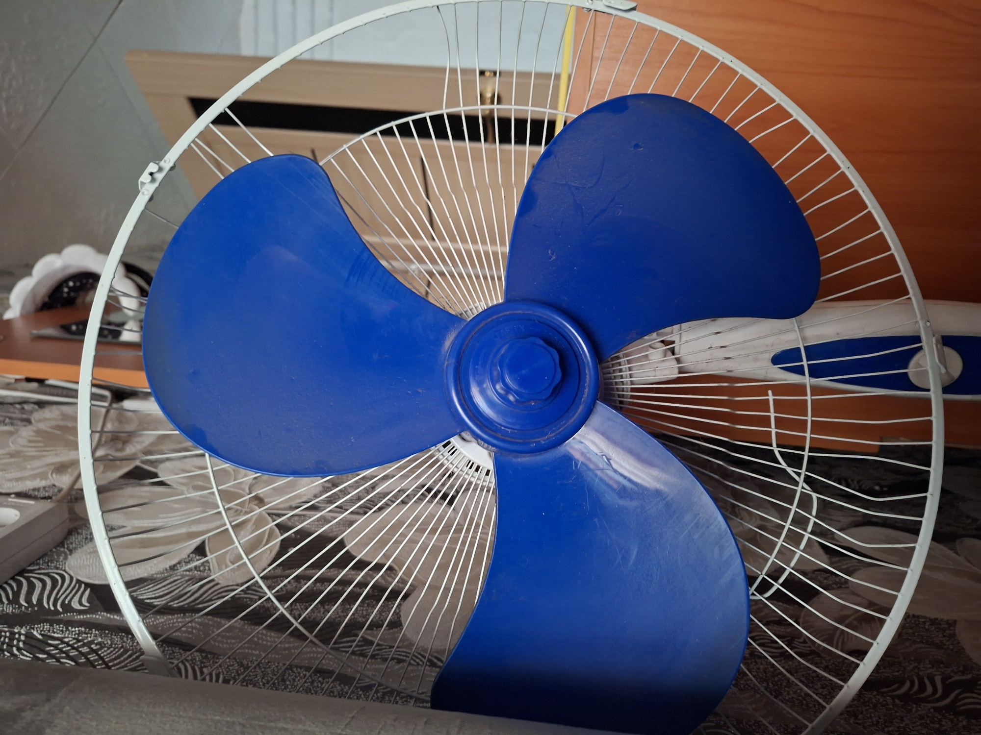 This is a close-up, eye-level, medium shot of a blue-bladed electric fan. The fan has a white metal grille and a white and blue motor housing. It is positioned in an indoor setting with a patterned wallpaper in the background, an orange wooden cabinet to the right, and a door visible further in the background. The image does not provide enough information to determine the specific location within the city of Amankaragay, Kazakhstan. The fan appears to be stationary.