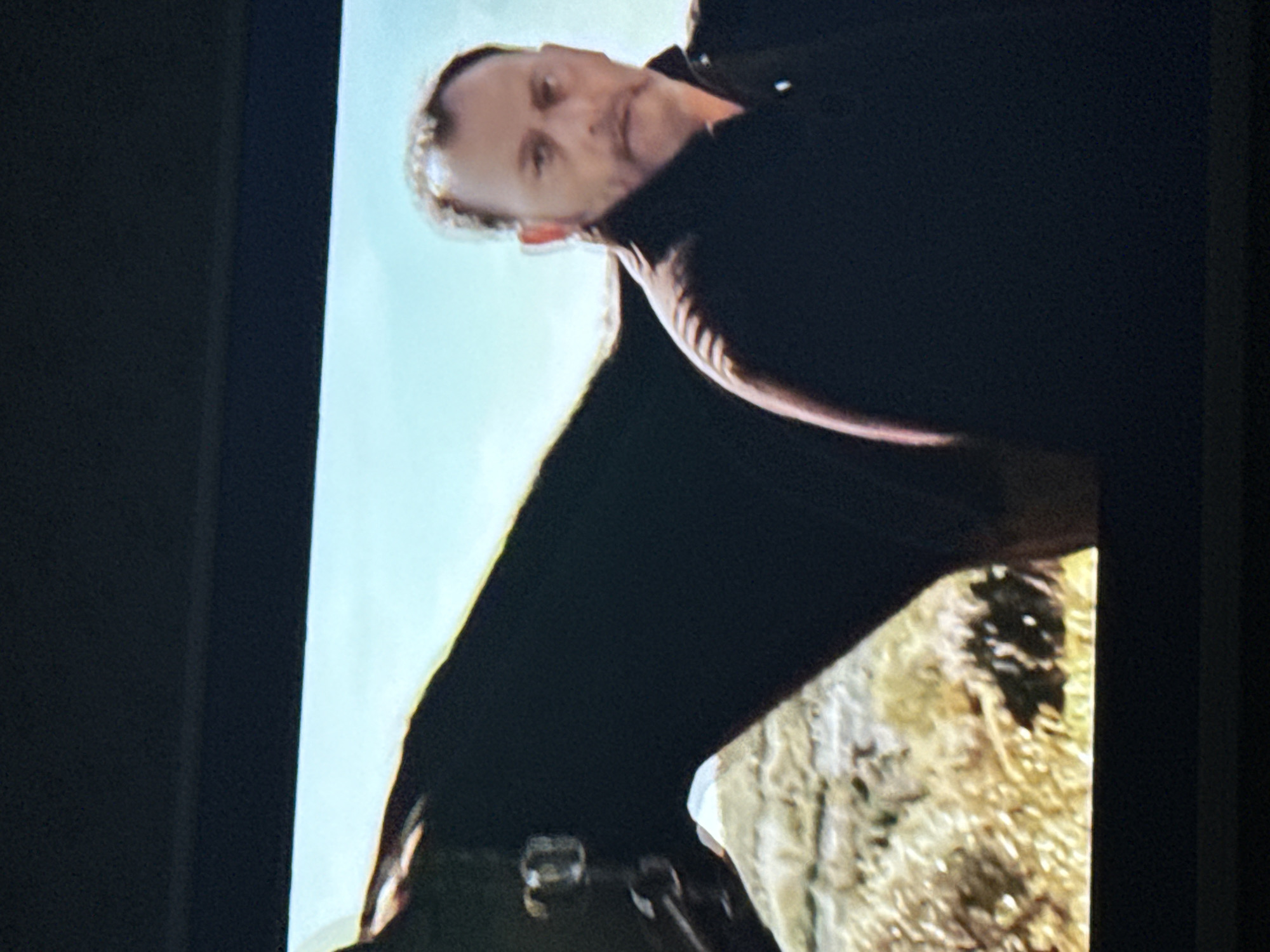 A man with short hair and a dark coat is riding a black horse. They appear to be in a field with tan and green vegetation. The sky is light blue.