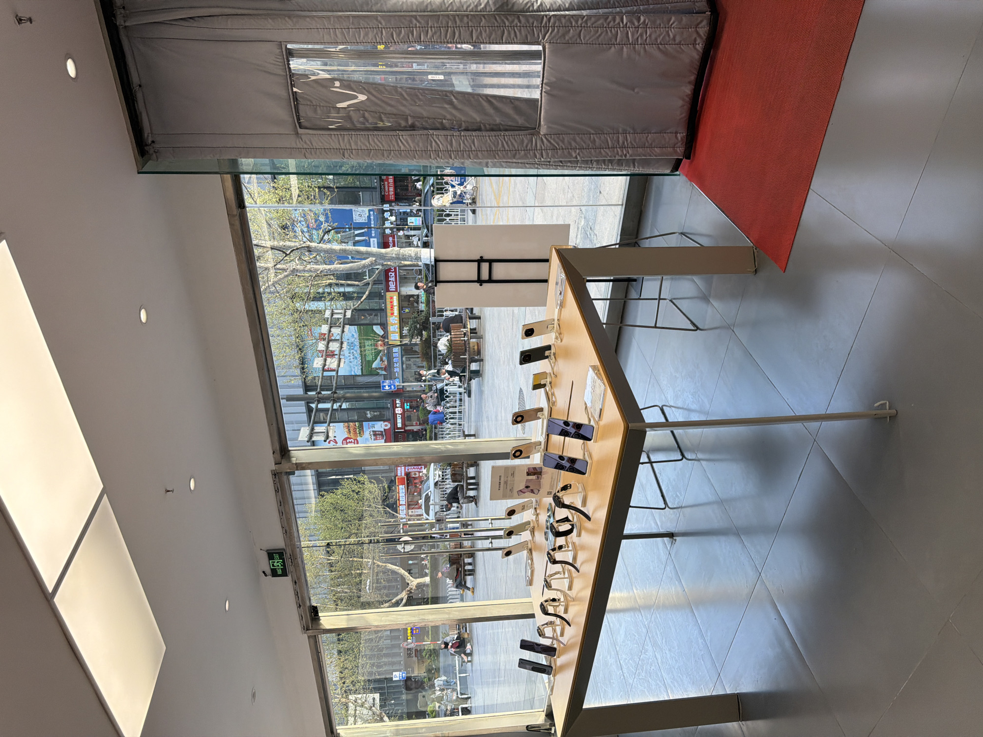The image depicts the interior of a store in Xuanwu District, China, looking out onto a busy city street. A table displays various electronic devices, including smartphones and smartwatches. The street scene visible through the large window reveals pedestrians, shops, and street signs with Chinese characters. The interior lighting is bright, with a modern ceiling and a red mat near a padded door.