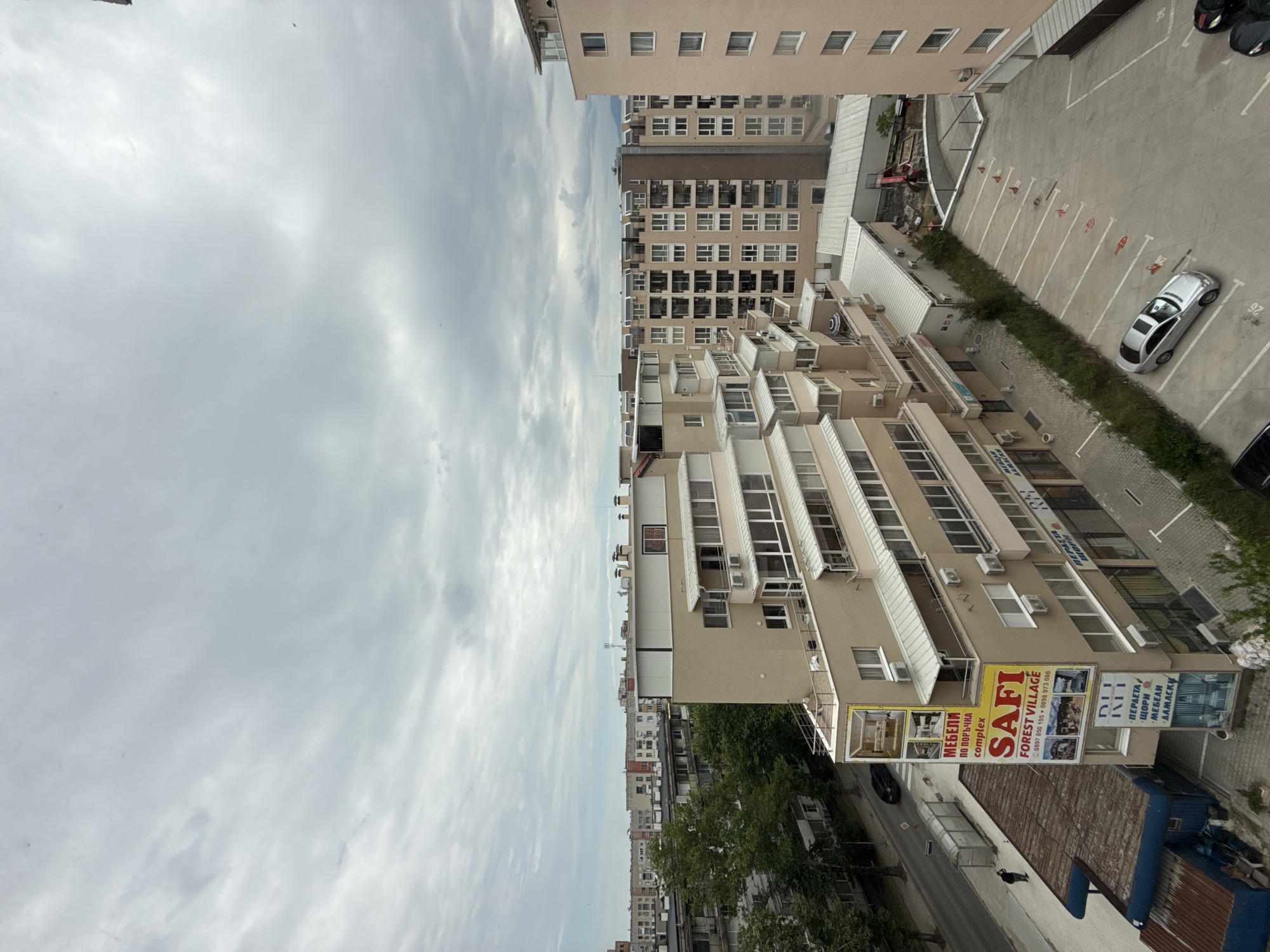This is an elevated view of an urban scene, likely taken from a high-rise building in Plovdiv, Bulgaria. The sky is predominantly cloudy. Several beige-colored buildings dominate the frame, some with rows of windows and air conditioning units. A parking lot is visible on the right side of the image, with marked parking spaces and a few cars, one of which is silver. A strip of greenery separates the parking area from the buildings. A street runs along the lower part of the frame, lined with trees and more buildings in the distance. A sign for "SAFI FOREST VILLAGE" is prominently displayed on one of the buildings, along with other Bulgarian text. There are no visible people in the scene.