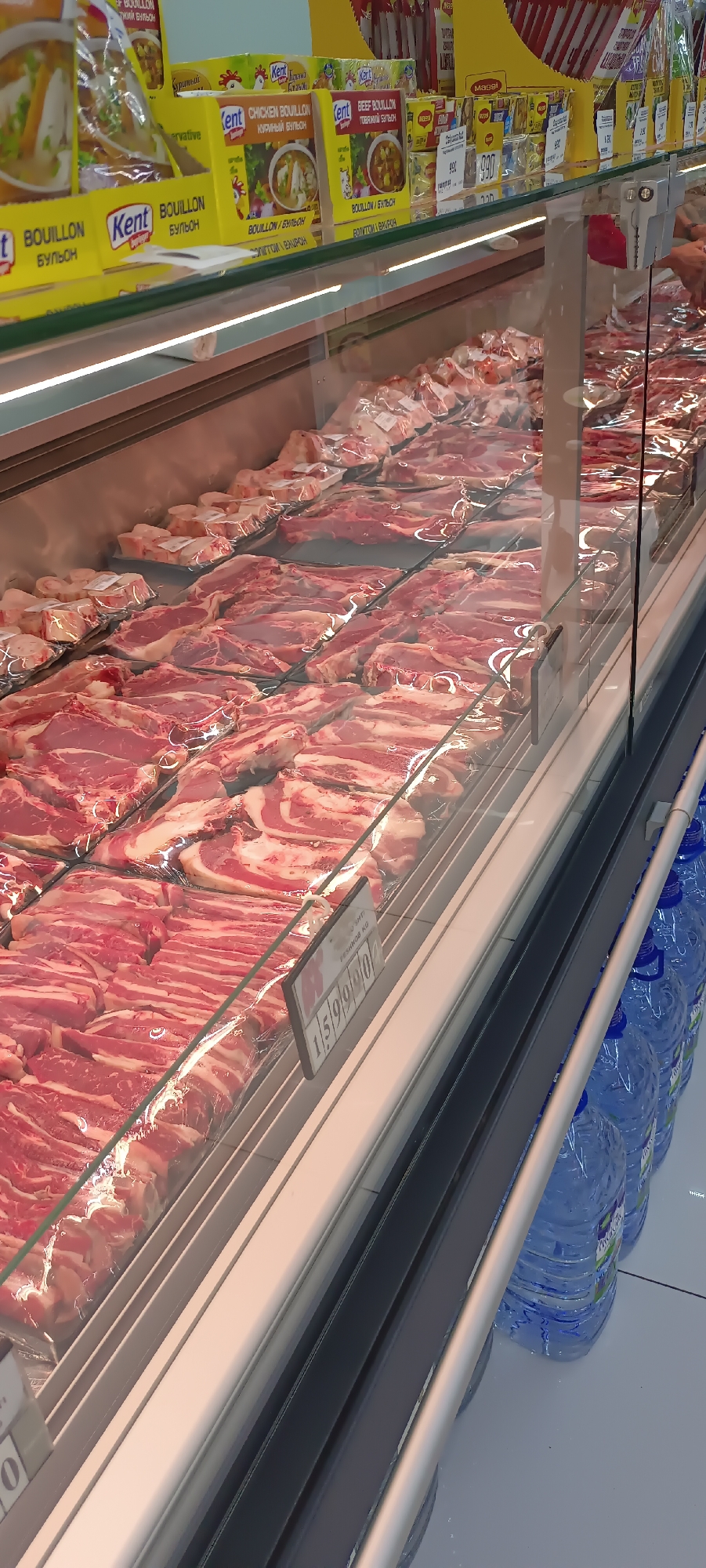 This media file shows a close-up view of a refrigerated display case filled with various cuts of raw meat, likely at a butcher shop or a grocery store in Fergana City, Uzbekistan. The meat is neatly arranged on shelves and appears to be fresh and well-packaged.
Above the meat display, several boxes of bouillon cubes are visible. Brands like "Kent" and "Maggi" are identifiable, with text in both English and Cyrillic scripts, suggesting a mix of international and local products. The text on the bouillon boxes includes terms like "Chicken Bouillon" and "Beef Bouillon," along with descriptions in Russian like "Куриный бульон" (Chicken Bouillon) and "Говяжий бульон" (Beef Bouillon).
A price tag is partially visible on one of the meat shelves, displaying a series of numbers: "1/5/9/9/0/0". The presence of this tag indicates that the meat is for sale, and this likely represents a price per unit of weight.
In the background, rows of large blue plastic bottles, possibly containing water, are lined up. The overall lighting suggests an indoor setting, likely during the day, as is common for retail environments. There are no people clearly visible in the frame, but the context implies a commercial setting where customers would be present. The image focuses on the merchandise, highlighting the variety of meat cuts and the associated food products.