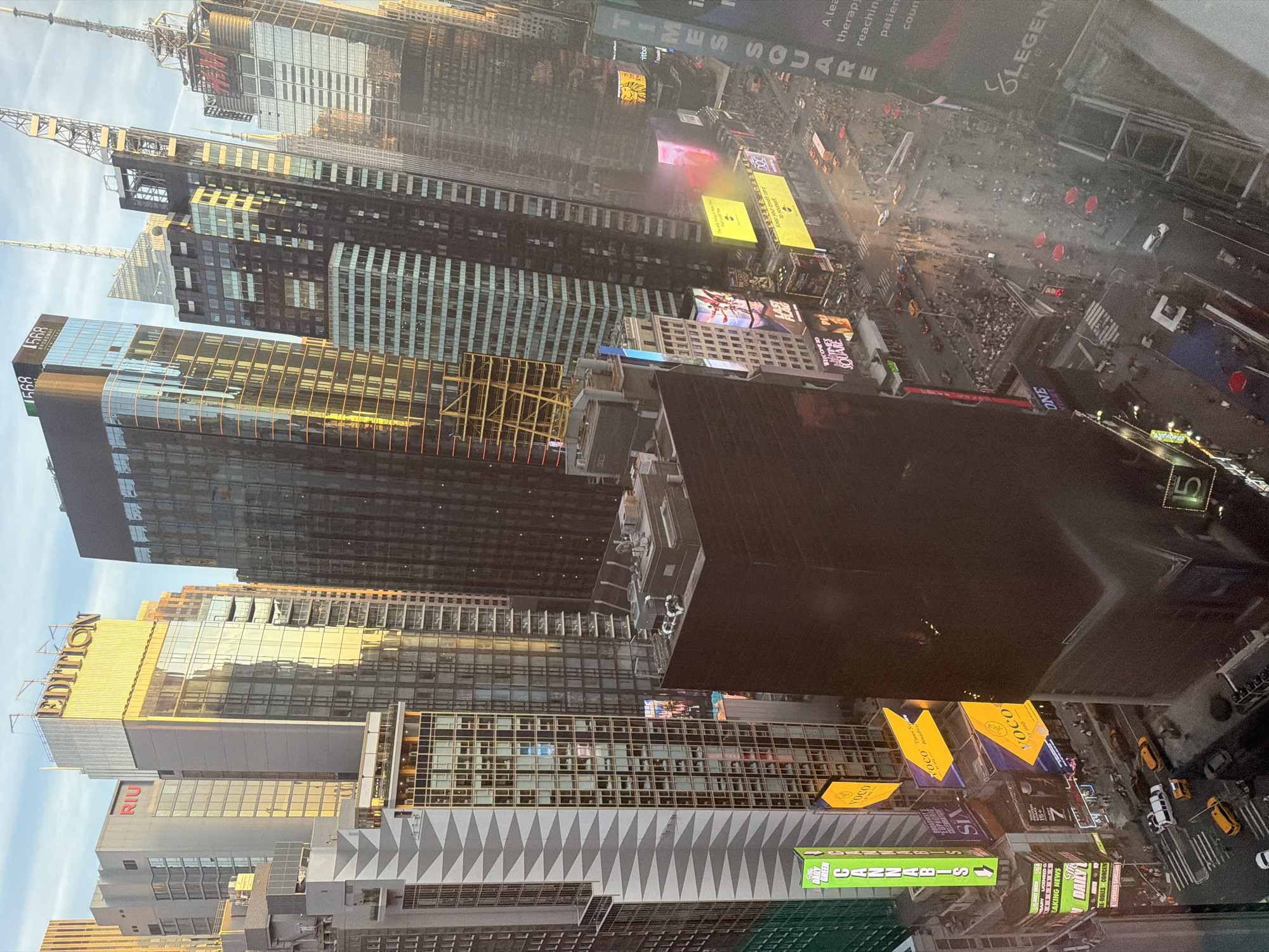 This aerial, slightly oblique view captures the bustling energy of Times Square in New York City, likely during late afternoon or early evening. Numerous towering skyscrapers, many with modern glass facades, fill the frame, reflecting a golden light on their upper sections, suggesting a setting or rising sun. Construction cranes are visible on some of the higher structures.
Below, the streets are alive with activity. Crowds of pedestrians are visible on sidewalks and crosswalks, while vehicles, including distinctive yellow taxis, navigate the thoroughfares. The scene is dominated by a multitude of massive, illuminated digital billboards and advertising screens that adorn the buildings, casting vibrant glows and creating a dynamic commercial landscape.
Notable details include the clear weather and the iconic density of billboards and traffic that immediately identify the location as Times Square. Visible text on buildings and digital displays includes "EDITION", "RIU", "568 BROADWAY", and "TIMES SQUARE". Advertisements feature brands like "CANNABIS", "DAILY GREEN", "VOCO", "SIX", and "LEGEND BIO". Other text snippets such as "DAILY BREAKING NEWS" and a partial message "KEEP YOUR CREDIT CARD TO YOURSELF" are also present, adding to the information density of the urban environment. The interplay of natural light on some buildings and the early illumination of the billboards highlights the transitional time of day.
