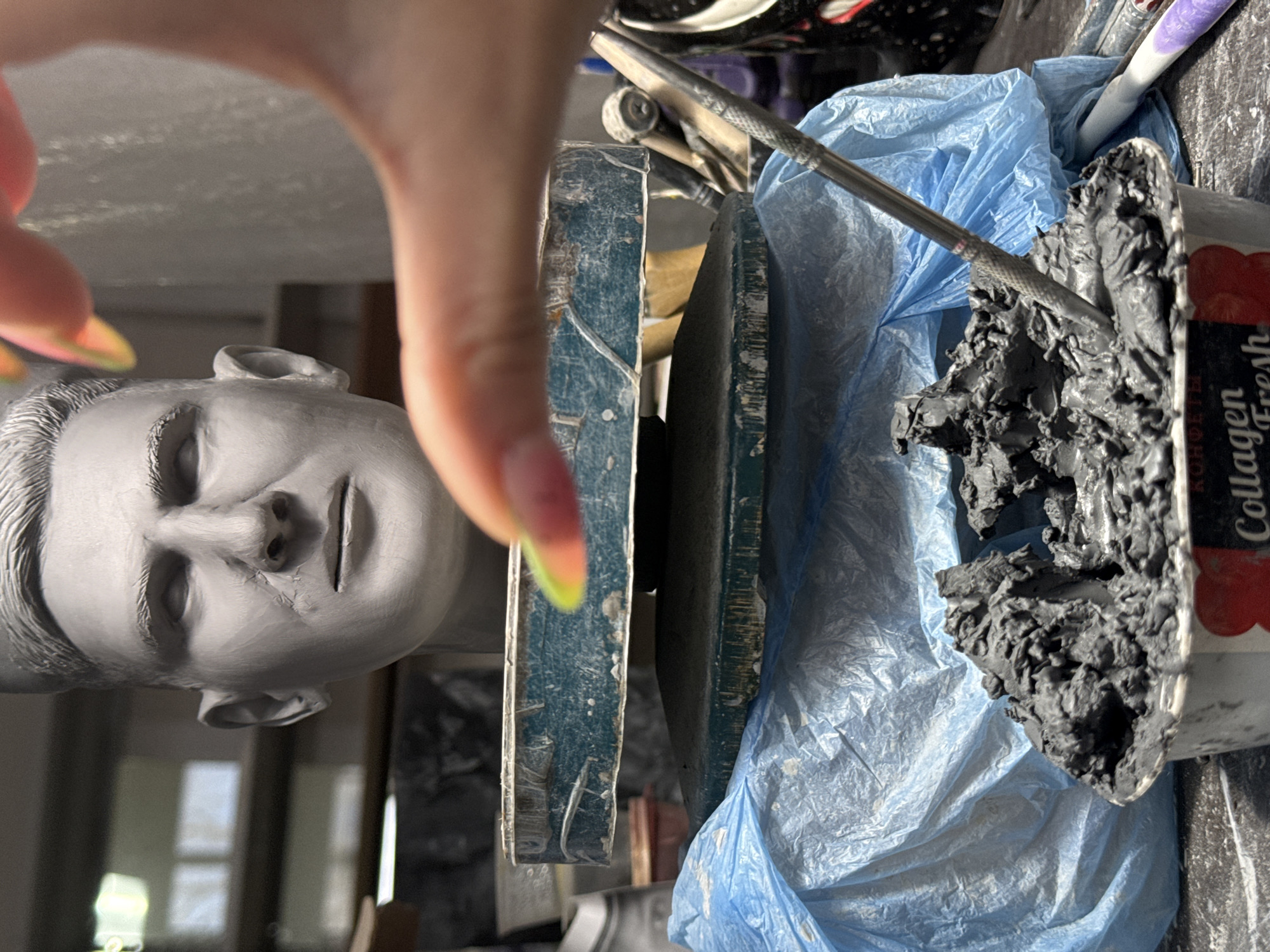 This vertically oriented image captures a detailed close-up of an art-making process, likely in a studio setting in Chaikovsky, Russia. In the foreground, a human hand with long, pointed nails, featuring a vibrant yellow, orange, and pink gradient manicure, extends from the top of the frame.
To the left, a grey clay sculpture of a human head, possibly a male bust, lies horizontally. The sculpture is finely detailed, depicting a serene face with closed eyes, a defined nose, lips, and sculpted hair. Its orientation suggests it is upside down relative to the hand.
In the center-right, a mound of dark grey, raw clay is held within a partially visible white and red container. A metal sculpting tool, featuring a textured handle, rests on this mass of clay. Beneath the clay and tool, a crinkled blue plastic bag is spread out, covering part of the workspace. Two worn, possibly wooden or metal, planks or boards are positioned horizontally across the mid-ground, acting as a surface or support structure for the sculpting materials.
The scene clearly depicts the activity of sculpting. The artist's hand, the partially finished sculpture, raw clay, and specialized tools all point to this creative endeavor. The bright, even lighting suggests an indoor environment, such as an art studio or workshop.
Visible text on the red and white container includes the words "Collagen" and "Fresh," with "KOROVKA" also partly discernible, indicating it is likely a repurposed candy box. The image conveys an intimate glimpse into the manual process of clay artistry.