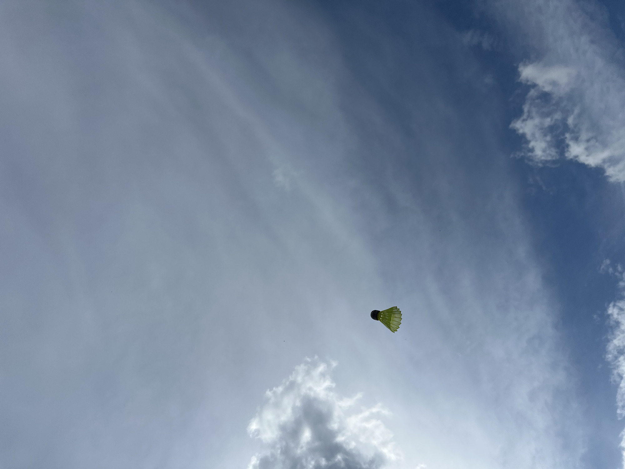 A badminton shuttlecock is suspended mid-air against a backdrop of a cloudy sky. The sky transitions from light gray clouds in the lower left to a more blue sky with white, patchy clouds on the right. The shuttlecock has a dark, possibly black, head and yellow feathers. There is nothing to indicate that the image was taken in Nizhny Novgorod or Russia.
