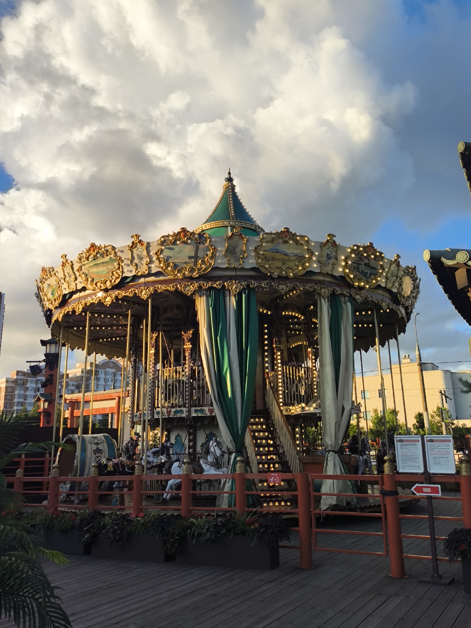 An outdoor carousel with a gold and green ornate canopy is the central focus of the image, set against a sky filled with dramatic clouds. The carousel features a detailed upper section with decorative panels and numerous lights, while green and white curtains hang down its sides. Inside, various carousel animals, including horses, are visible, along with stairs leading up to the ride. The carousel is surrounded by a red railing and planters filled with dark green foliage. In the background, there are multi-story buildings and trees. A wooden boardwalk or deck is in the foreground, leading towards the carousel. A sign with a red arrow and Russian text is also visible on a stand near the railing. The lighting suggests it is either early morning or late afternoon, with the sun casting a warm glow on the carousel.