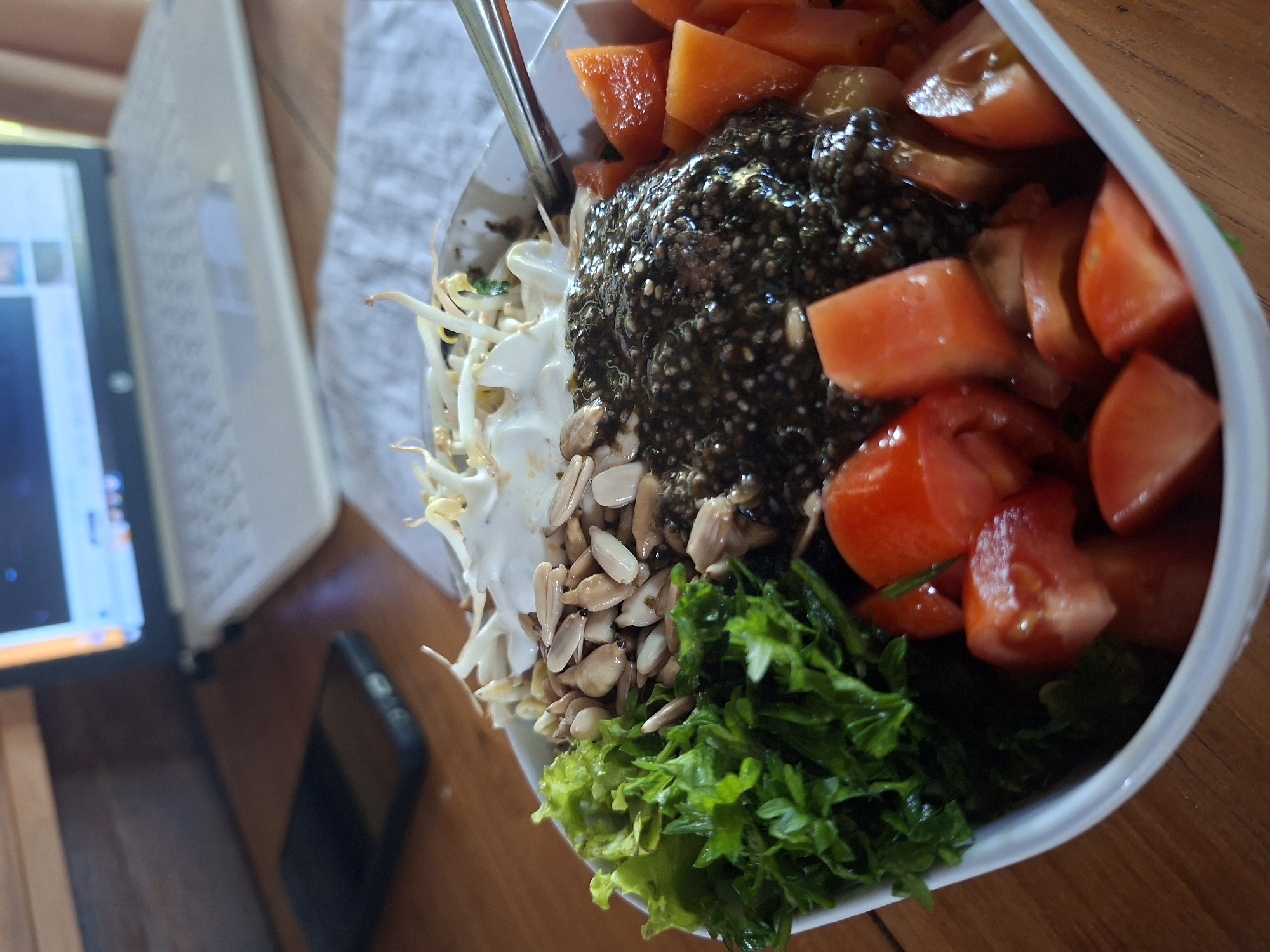 A close-up shot depicts a salad bowl, featuring tomatoes, papaya, a dark, seedy substance, sprouts with a white sauce, roasted nuts, and leafy greens. A laptop and phone are situated beside the bowl on a wooden surface. The location is Lodtunduh, Indonesia.
