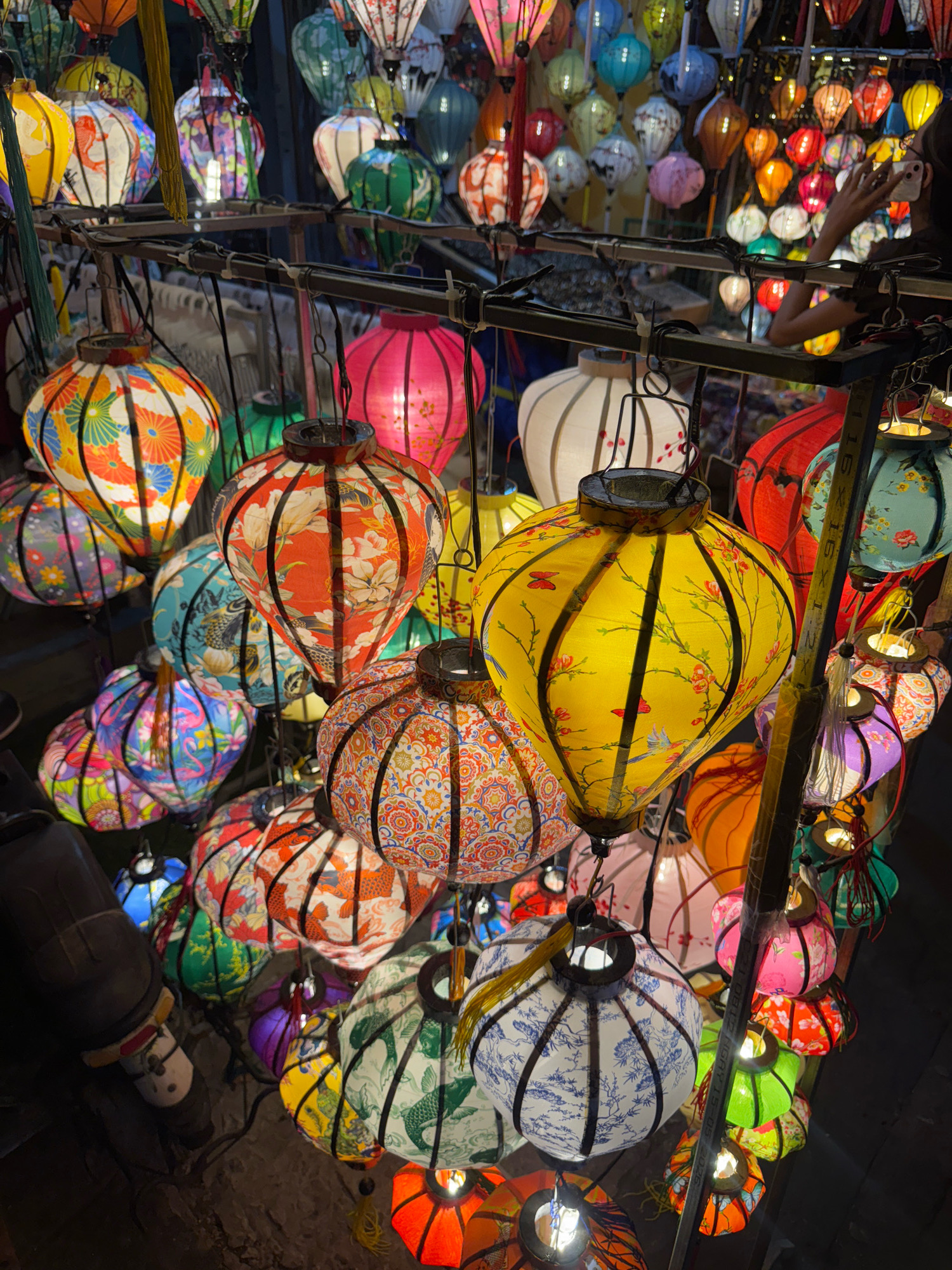 This is an outdoor night scene in Hoi An, Vietnam, featuring a dense display of colorful, patterned fabric lanterns. The lanterns are of various shapes and sizes, predominantly spherical or oval, and are illuminated from within, casting a warm glow. They are hung from metal racks and wires, creating a vibrant and somewhat cluttered visual landscape. The patterns on the lanterns include floral designs, birds, fish, and abstract motifs in a wide spectrum of colors, such as red, yellow, blue, green, pink, and orange. In the background, a person is partially visible, holding a phone and appearing to take a photo, indicating a tourist or local engaging with the scene. The overall impression is one of a lively marketplace or cultural attraction, characteristic of Hoi An's nighttime ambiance.