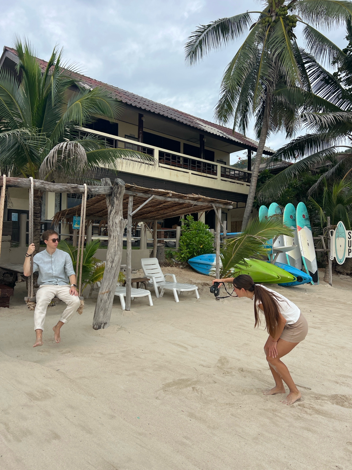 A young man in a light blue striped shirt and off-white pants sits on a wooden swing on a sandy beach. He is wearing sunglasses and has a watch on his left wrist. To his right, a young woman in a white t-shirt and beige shorts is crouched down, taking a photo with a black camera. In the background, there is a two-story building with a tiled roof, surrounded by palm trees. Several kayaks and stand-up paddleboards are propped up against the building. Two white plastic lounge chairs are also visible on the sand. The scene appears to be a resort or guesthouse in the Ko Samui District of Thailand. The sky is overcast.