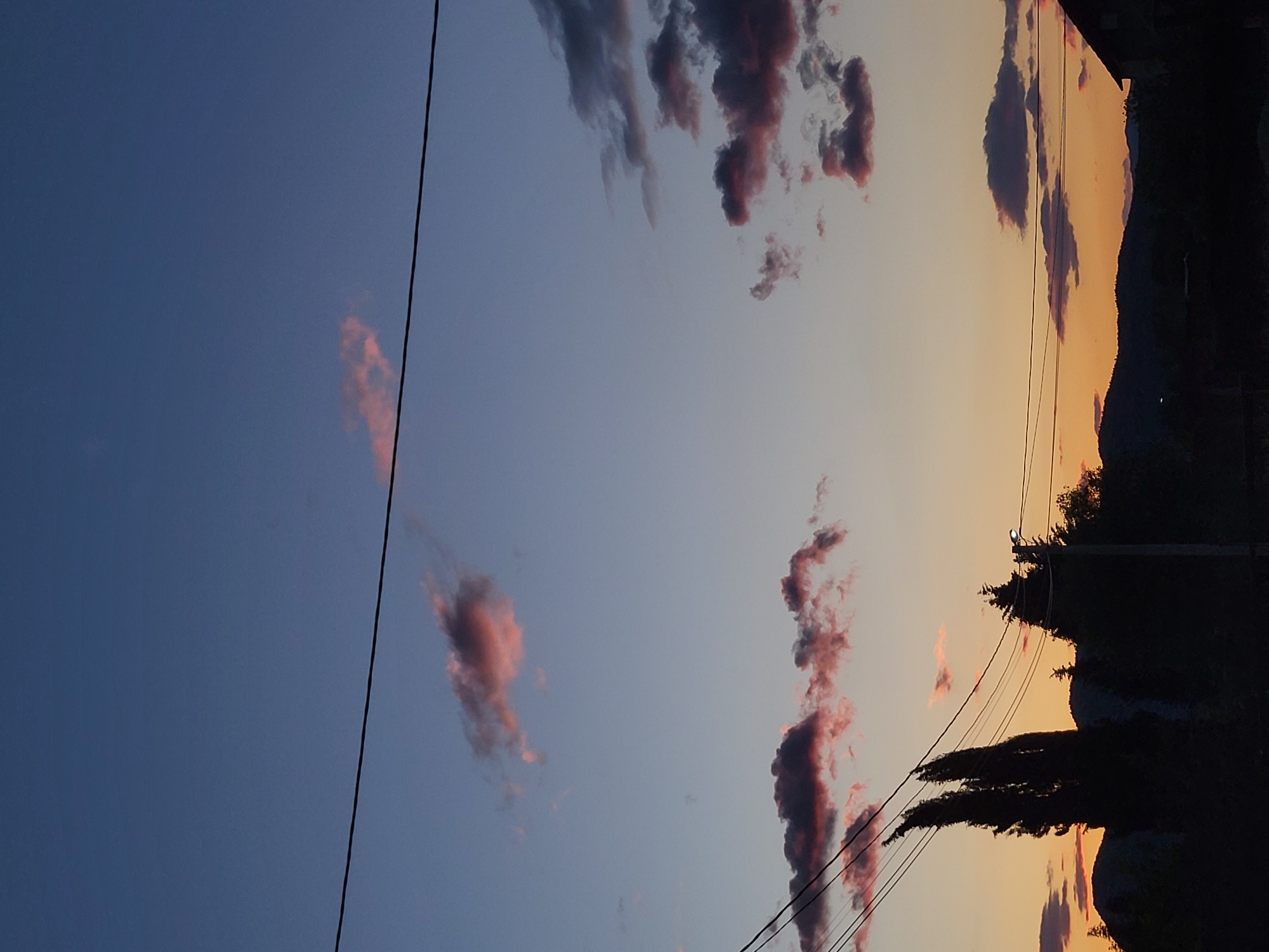 The image, taken in Mahala, Montenegro, depicts a serene sunset or twilight scene. The sky transitions horizontally from a deep blue on the left to warm orange and yellow hues on the right. Scattered clouds are visible across the sky; some are dark grey and purple, while others are illuminated with soft pink and reddish tones by the setting sun.

Several dark, thin power lines stretch diagonally across the sky from the upper left towards the lower right. On the right side of the image, the silhouette of mountains or hills defines the horizon, set against the brighter portion of the sky. Below these mountains, dense, dark silhouettes of trees and other vegetation are prominent in the foreground. A faint, dark outline of what appears to be a corner of a building or structure is barely discernible on the far right, above the treeline.

The time of day is clearly evening, either sunset or twilight, due to the color gradient of the sky and the silhouetted landscape elements. The weather appears to be partially cloudy. No text is visible in the image.