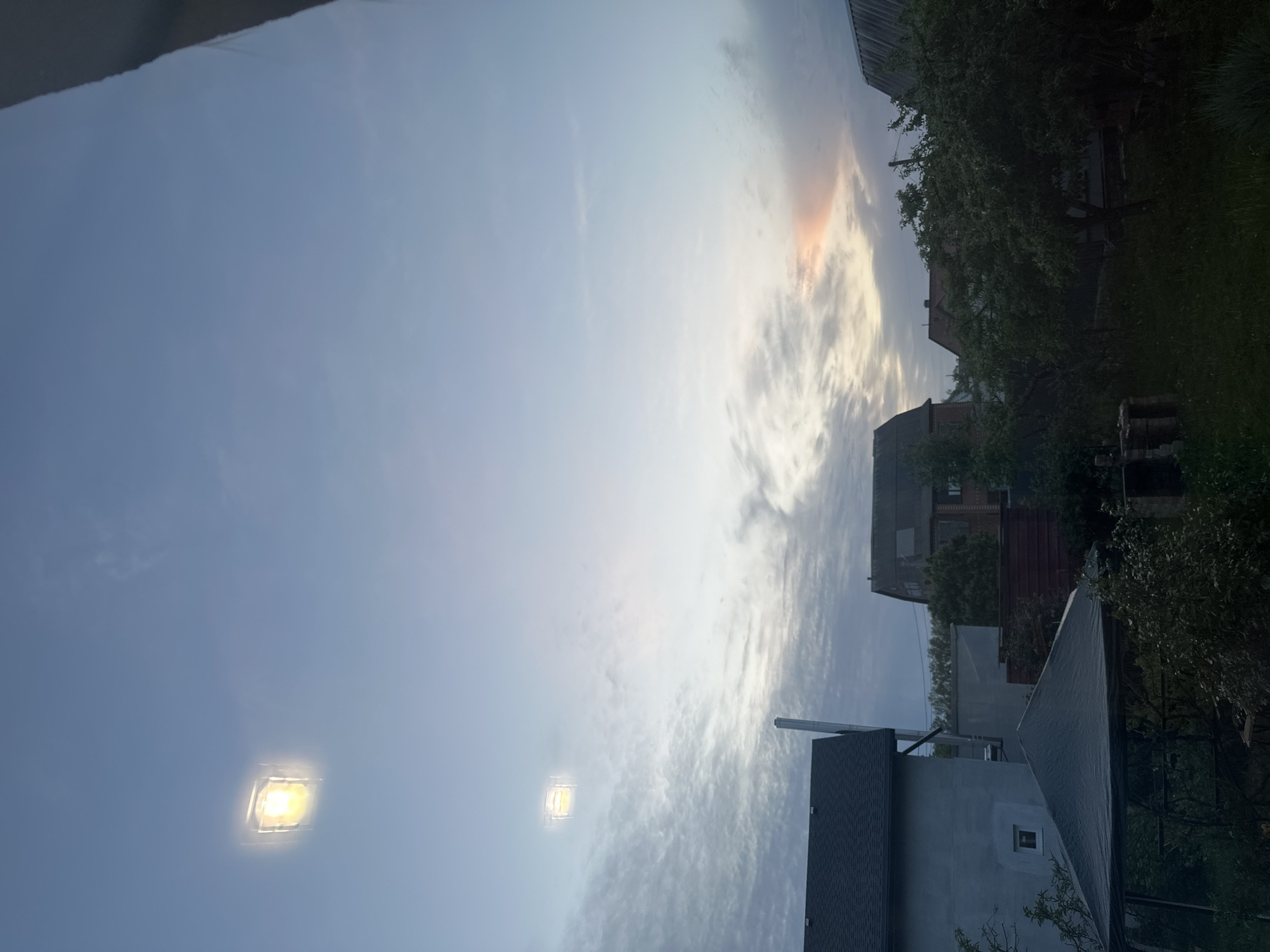 The image shows a view looking up towards the sky in Machukhy, Ukraine. The sky is predominantly blue with scattered clouds, some tinted with orange hues. There are buildings and trees visible on the right side of the frame. Two bright, square-shaped lights are visible in the sky.