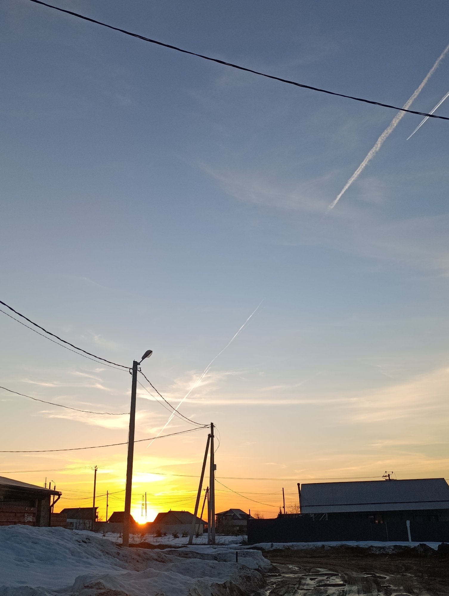 The image shows a street scene at sunset in what appears to be a residential area. Patches of snow cover the ground alongside muddy areas with puddles. Several houses and a large building are visible, some with dark fences. Electrical poles and wires crisscross the sky. The sun is setting behind the houses, casting a warm orange glow. Vapor trails from aircraft are visible in the sky. The location is potentially Chaikovsky, Russia. No people are visible in the image.