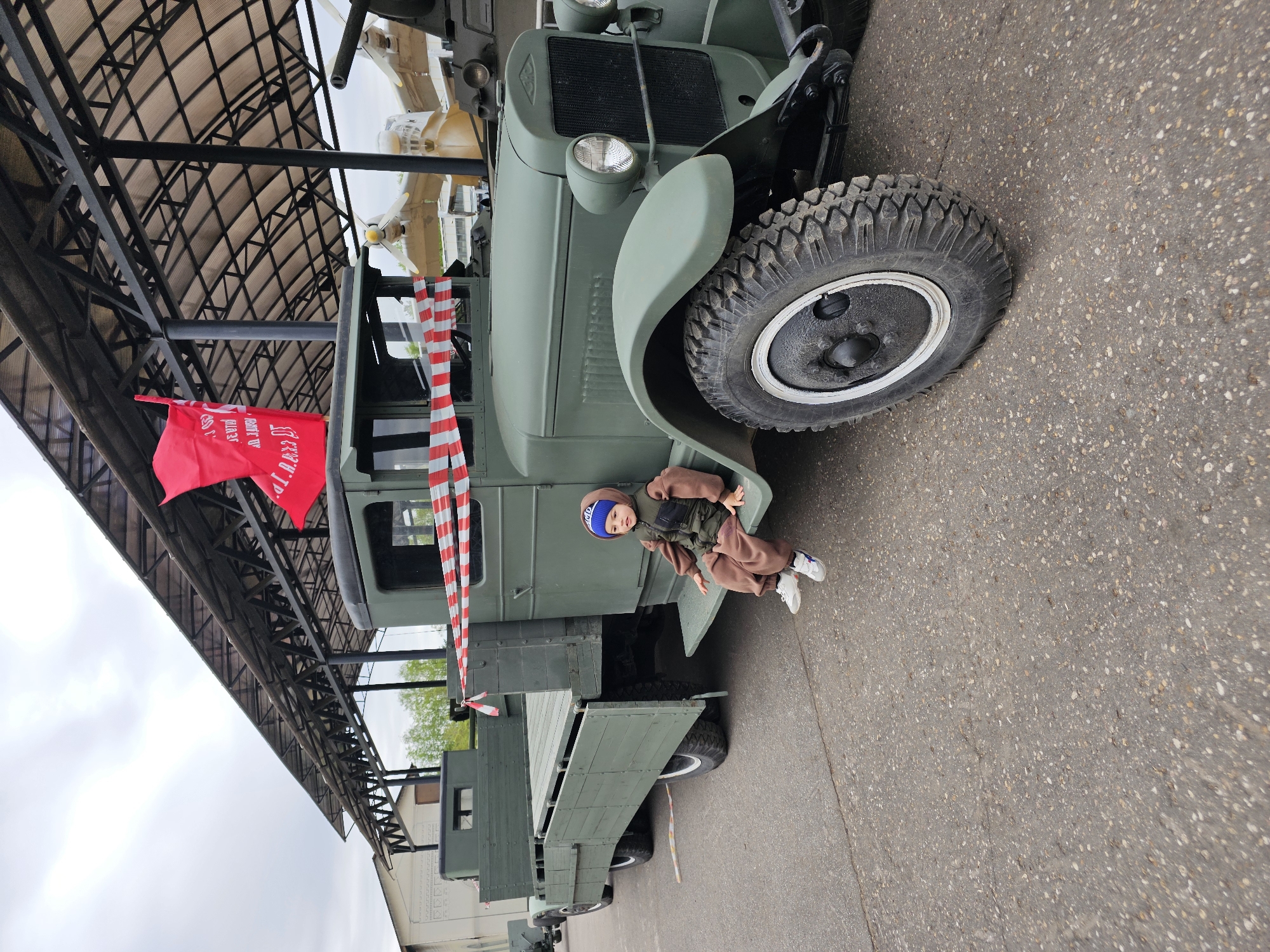 The scene shows a green military truck with a child sitting on its fender, parked outdoors under a metal structure. The truck has a red flag and red and white striped ribbon attached. The child wears a blue hat, brown jacket and pants, and white sneakers. The location is Arkhangel’skoye, Russia. The ground is asphalt.