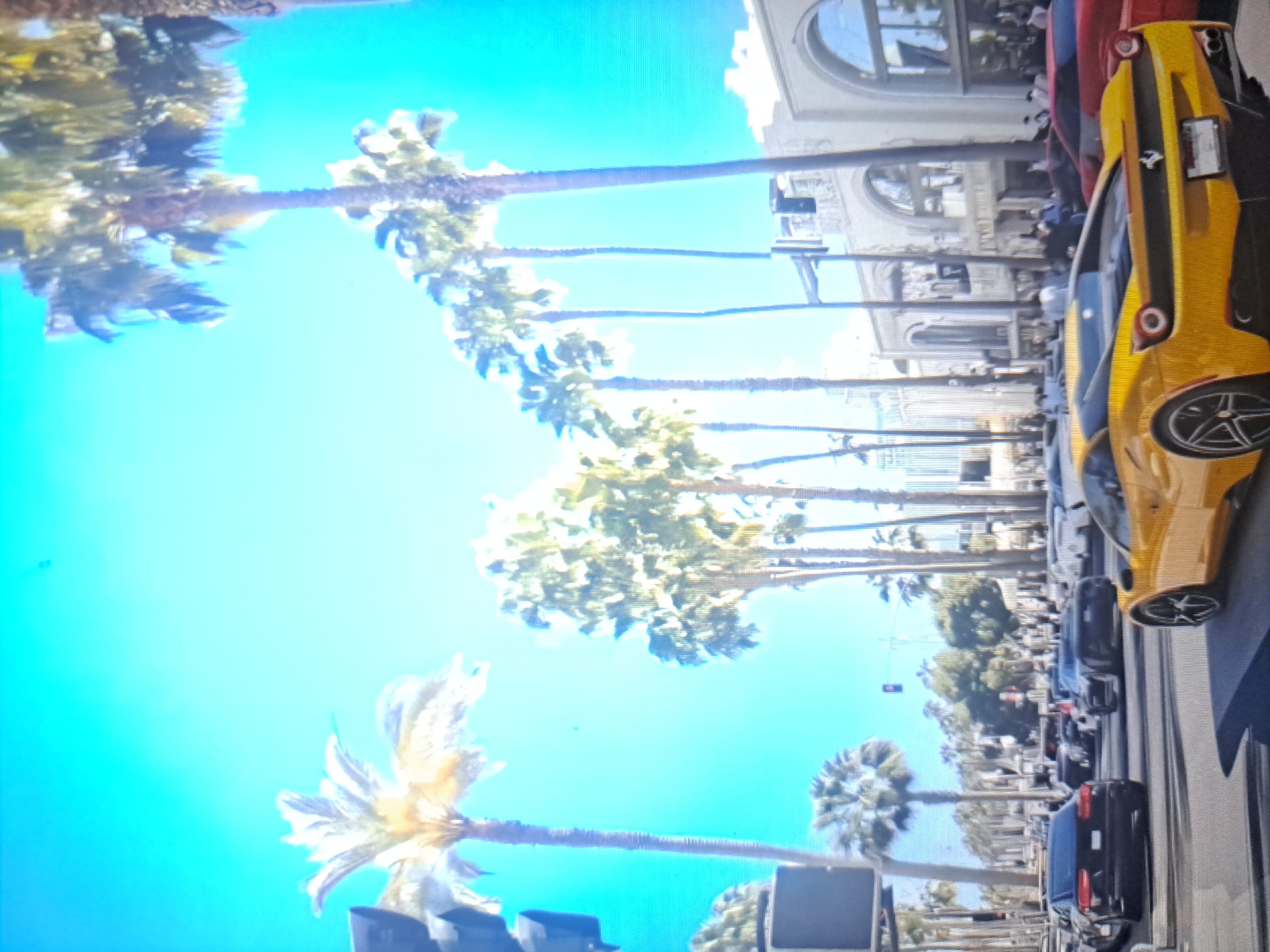 The image depicts a street scene with a yellow sports car parked on the right side of the road. Palm trees line the street, suggesting a tropical or sub-tropical location. Buildings are visible in the background, hinting at an urban environment. A row of cars is parked along the street, indicating a busy area. The image appears to be taken on a clear, sunny day. The location is not Yerevan, Armenia, but rather a coastal region with palm trees.
