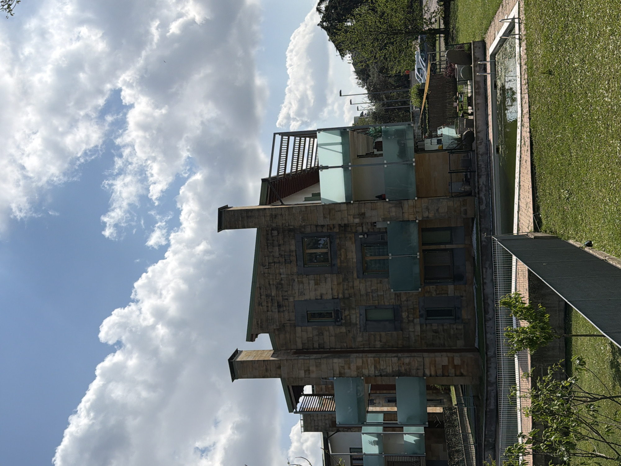 The image presents an outdoor scene featuring a multi-story building, a garden, and a swimming pool under a bright, partly cloudy sky, likely set in Vigevano, Italy.
The building, constructed with stacked stone or brick, showcases a modern design with numerous windows and balconies. These balconies are equipped with frosted or light-colored glass railings and accented by wooden slatted overhead structures. Two prominent, stone-clad chimney-like elements rise from the roofline.
A well-kept green lawn stretches beside the building, encompassing a rectangular swimming pool. The pool water appears somewhat murky or green, suggesting it may not be in active use or recently maintained. Steps or a ladder are visible within the pool at one end.
The backdrop includes a line of dense green trees, while a few branches extend into the foreground, contributing to a lush environment. The sky is bright blue, dotted with large, fluffy white clouds, indicating a sunny day, likely midday or early afternoon. A section of a road or parking area, with a white vehicle, is faintly discernible in the distance. Outdoor furniture is subtly visible near the pool. A fence delineates parts of the property.
No people or explicit activities are depicted, giving the scene a tranquil and serene atmosphere. No visible text is present in the image.