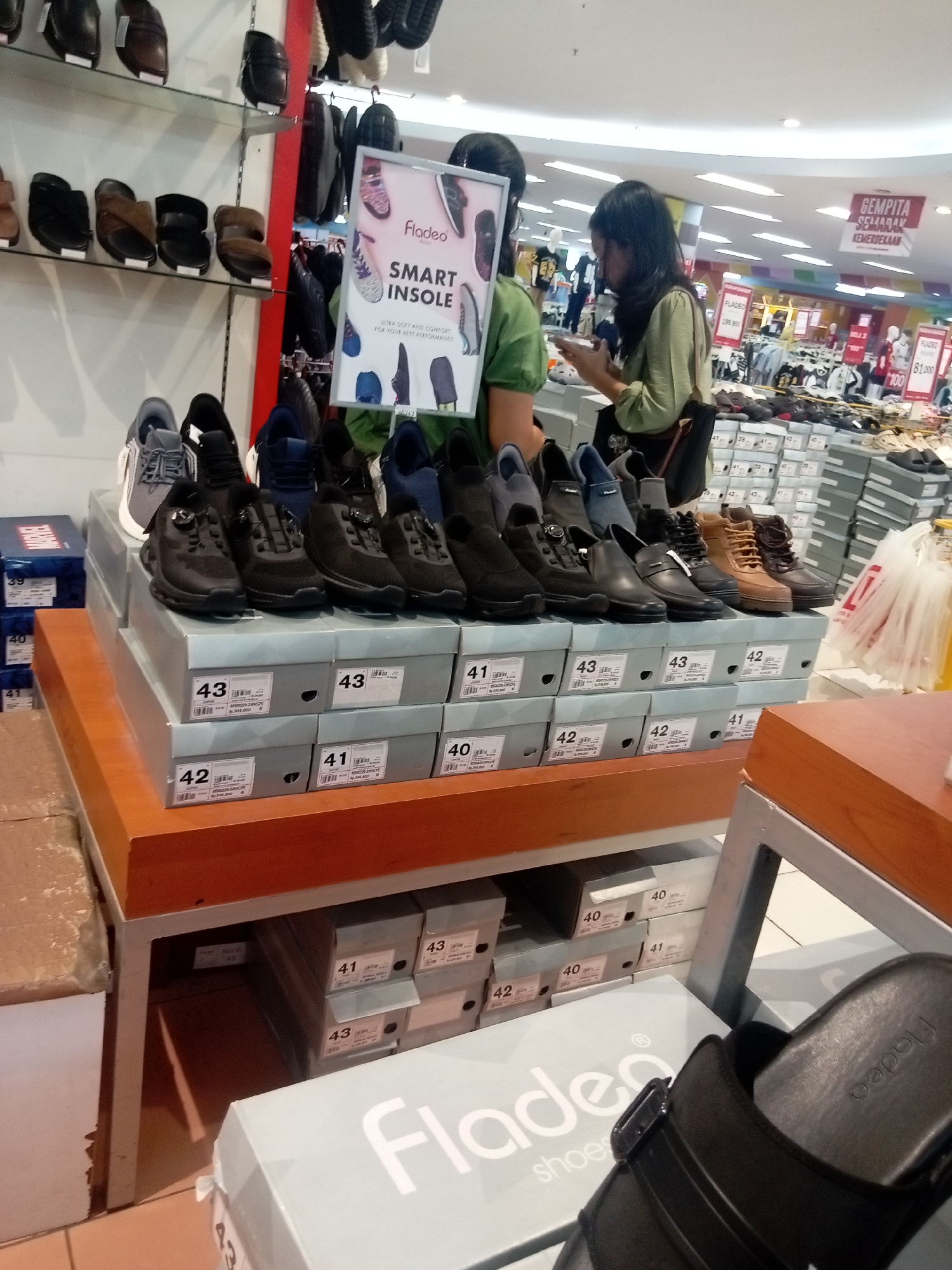 The image shows a shoe store interior, likely in Batam, Indonesia, with shelves and display areas filled with footwear. In the foreground, stacks of shoeboxes are organized on a wooden display table. Some shoeboxes have size labels such as "43," "41," and "40," along with visible prices. A black sandal with the brand name "Fladeo" is prominently displayed on a light blue shoebox in the bottom right corner.
Several pairs of shoes are displayed on top of the shoebox stacks, including black sneakers, boots, and casual shoes. In the mid-ground, a woman with long black hair, wearing a light green long-sleeved shirt, is looking at her phone while standing next to a display of shoes. Another person, partially visible, is wearing a green shirt and standing near a "Fladeo SMART INSOLE" advertisement.
The background reveals more shelves stocked with shoes and the retail environment of a store, including lighting fixtures and signage. One sign in red lettering reads "GEMPITA SEMARAK KEMERDEKAAN," suggesting a patriotic or festive sale event, common in Indonesia. The overall scene depicts a typical retail environment for a shoe store.