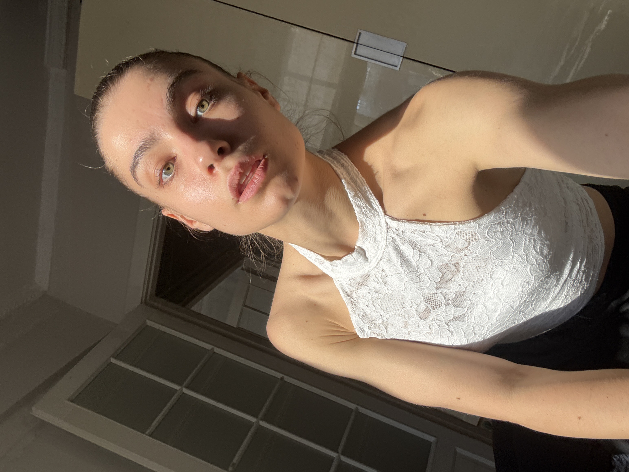 A light-skinned woman is angled from the lower left corner of the frame towards the upper right corner. She wears a white lace halter top and black pants. The light is strong on the upper left of her face, torso, and arm, casting shadows elsewhere. In the background, a window with a grid pattern is on the lower left, and a glossy white surface, possibly a cabinet, is on the upper right. The location is not identifiable as Moscow, Russia, from the visual elements of the image.