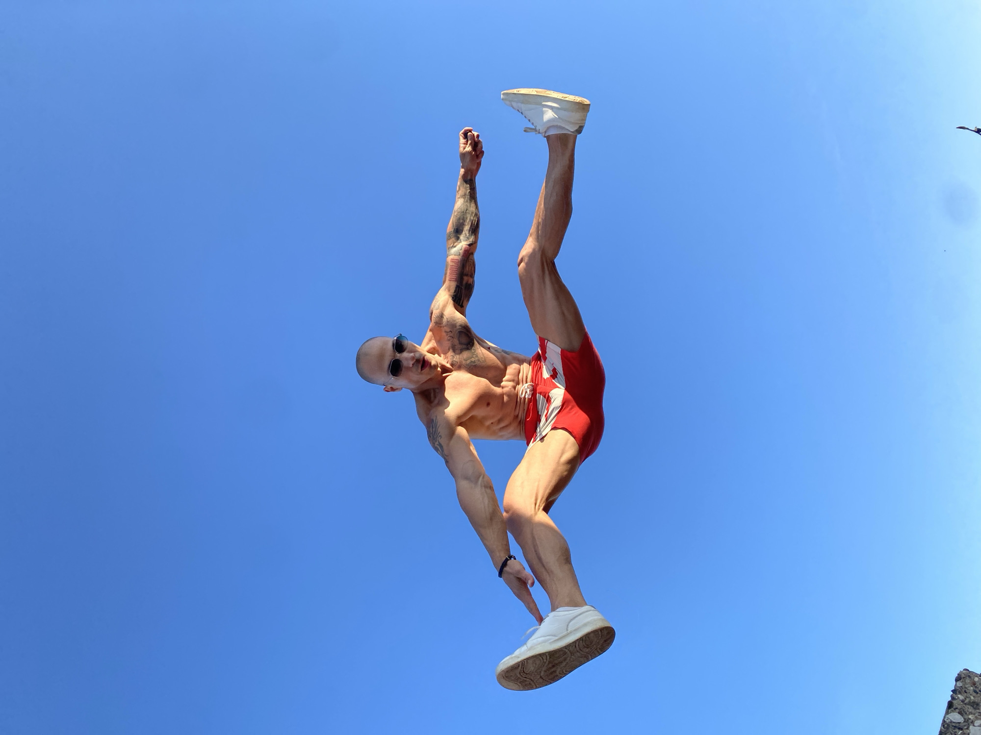 A shirtless, bald man with sunglasses and tattoos is captured mid-air, performing a split jump against a clear blue sky. He is wearing red and white patterned shorts and white sneakers. His body is oriented horizontally, with his legs extended outwards and his arms outstretched, mirroring the split. The background is a uniform, bright blue sky with no visible clouds or objects, suggesting an outdoor setting likely during the daytime. The perspective is from below, looking up at the man. The lower left corner shows a small portion of a stone structure.