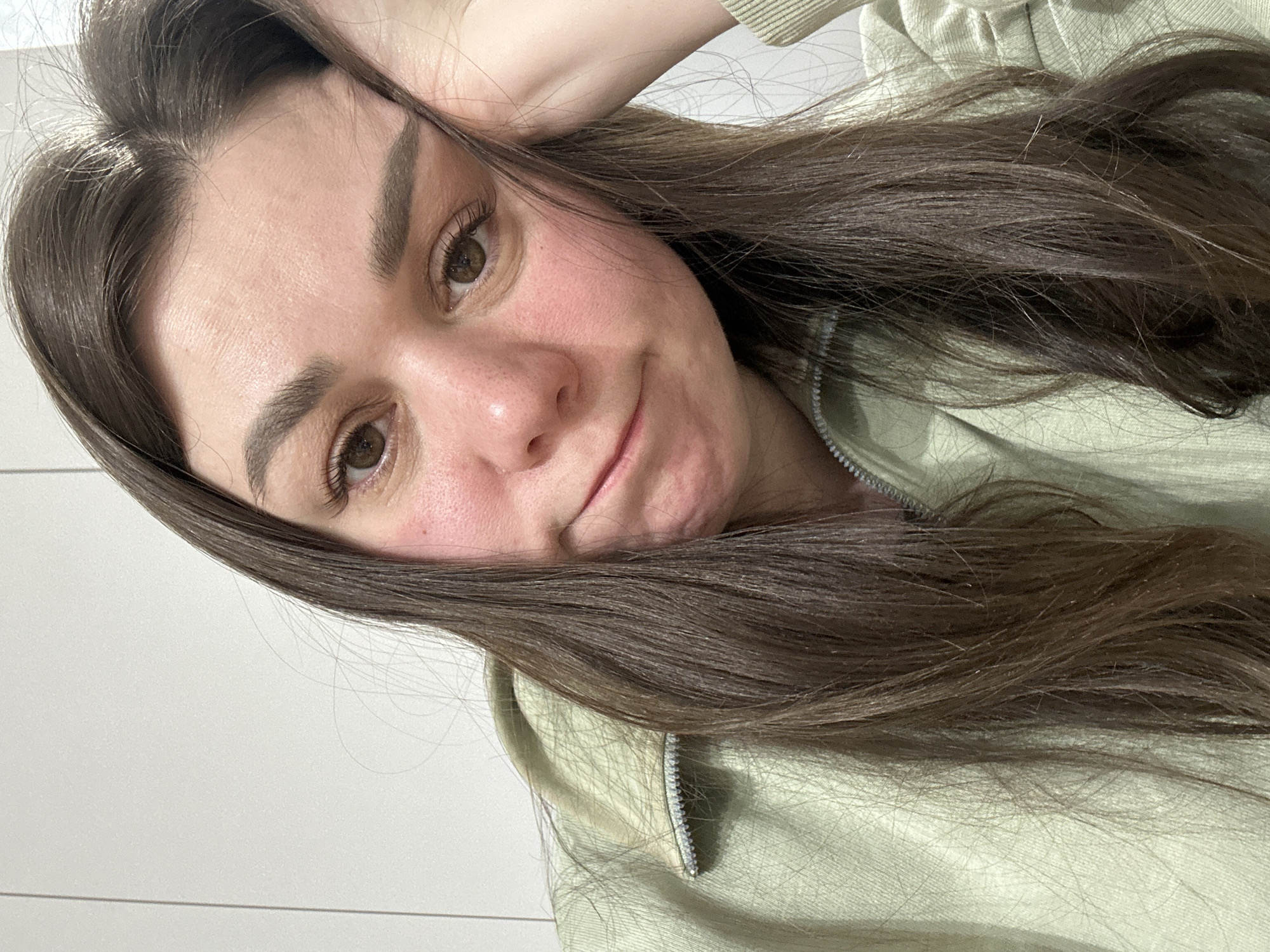 A close-up, angled portrait features a young woman with long, dark brown hair and brown eyes, likely taken indoors in Chaikovsky, Russia. Her head is tilted, and her right hand is gently placed near her ear, partially obscured by her hair. She looks towards the camera with a subtle, somewhat pensive or slightly amused expression, her lips forming a small, soft curve.
Her complexion shows a natural texture with visible pores and a slight flush on her cheeks. Her eyebrows are well-defined, and her eyelashes are visible. She is wearing a light sage or mint green zippered top, possibly a hoodie or jacket, which drapes over her shoulder. Her dark hair flows down, covering parts of her face and the left side of the frame, with individual strands visible.
The background is a plain, light-colored wall, possibly white or off-white, with faint horizontal lines that suggest paneling or wallpaper. The lighting appears soft and even, typical of indoor settings. No specific time of day or weather conditions are discernible. There is no visible text in the image.