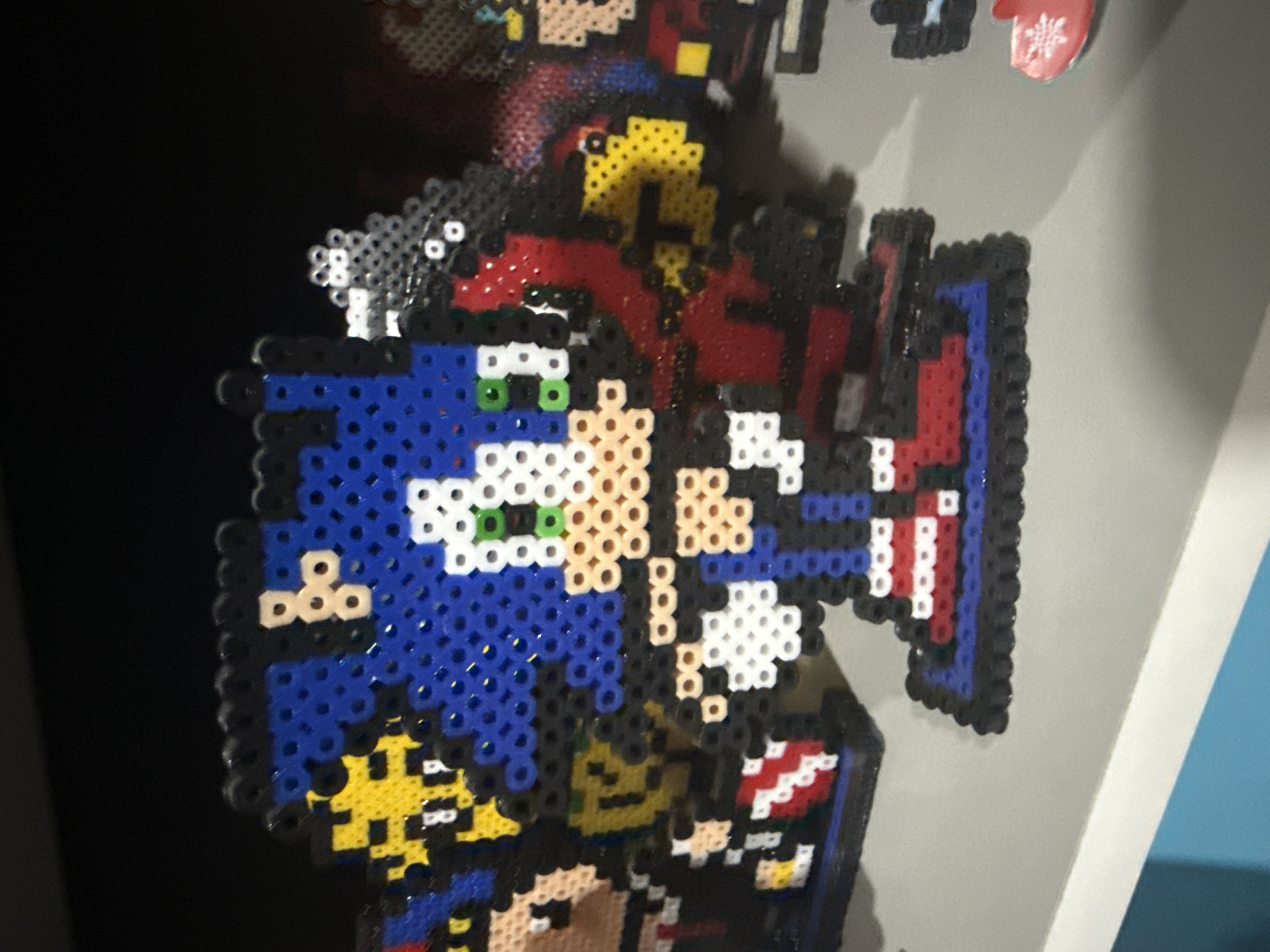 The image presents a close-up view of several perler bead artworks, likely displayed on a vertical surface indoors. The central and most prominent piece features a unique split design combining two distinct characters. On the left, Sonic the Hedgehog is visible with his iconic blue quills, peach-colored face, and bright green eyes. On the right, Captain America is depicted, recognizable by his signature red, white, and blue shield with a central star, along with segments of his red and blue uniform.
Surrounding this main artwork, other perler bead creations are partially visible. Above the central piece, part of a character with yellow and red elements, possibly Iron Man, can be seen. In the upper left, hints of other pixelated figures are present but too indistinct for clear identification. To the top right, a small, red and white mitten-shaped sticker or magnet featuring a white snowflake design is visible.
The artworks are crafted from numerous fused plastic beads, showcasing vibrant colors including blue, red, white, yellow, green, black, and peach. The pixelated aesthetic characteristic of perler bead art is evident throughout. The scene is illuminated by what appears to be indoor lighting, with a dark background on the left and a lighter, possibly white or gray surface on the right. No discernible text is present in the image. This display of creative perler bead art is situated in Puebla, Mexico.