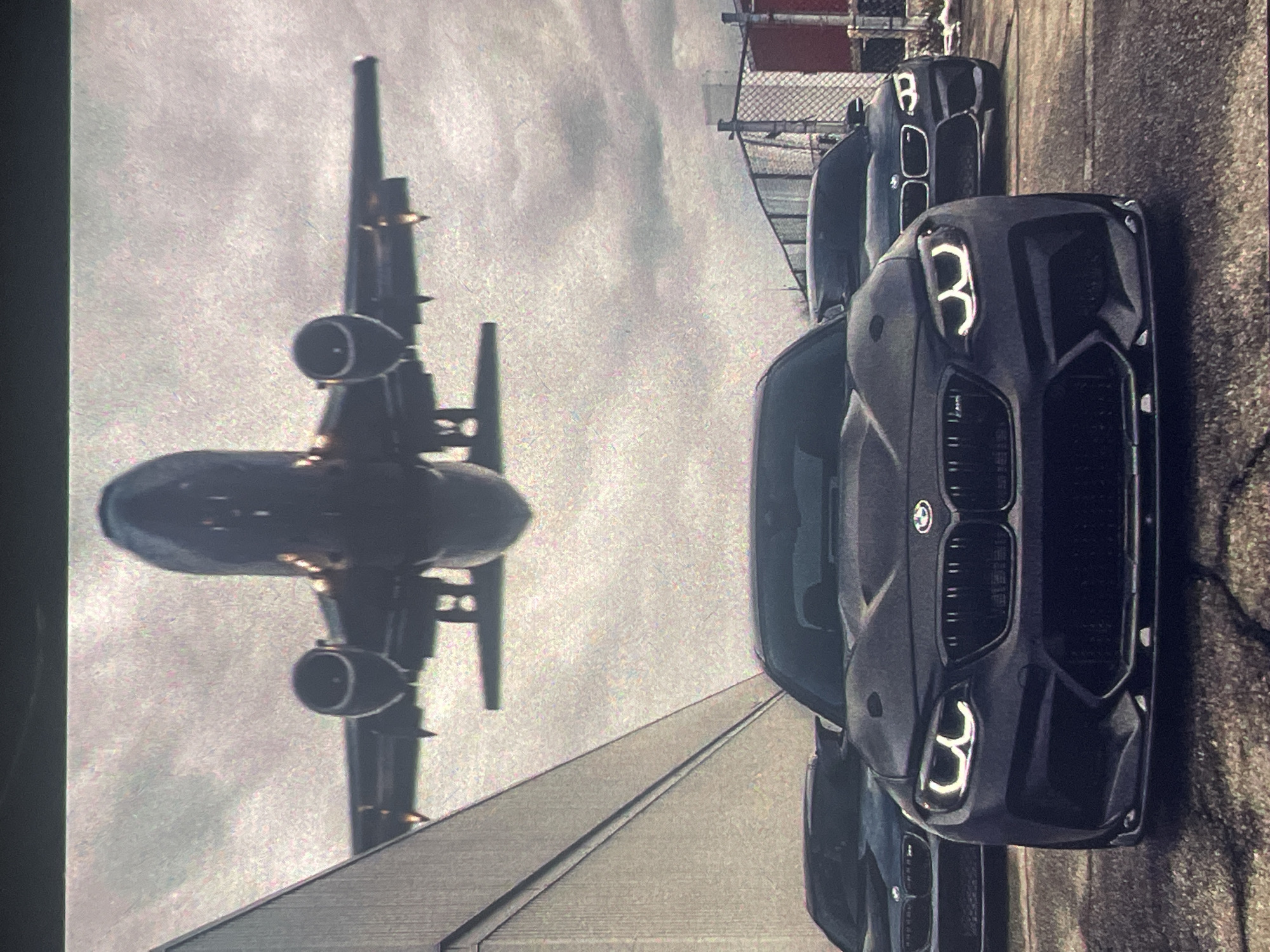 An airplane is captured in mid-flight, descending towards a runway. Below the aircraft, three dark-colored BMW cars are parked on an asphalt surface. The car in the foreground is a dark gray BMW M2, prominently displaying its distinctive headlights and front grille. To the left and right of the M2, two other BMWs are visible, partially obscured. The environment appears to be an industrial or airport setting, with a large building to the left and a chain-link fence in the background. The sky is overcast, suggesting a cloudy day. The scene does not contain any people or specific information about Paczkowo, Poland.