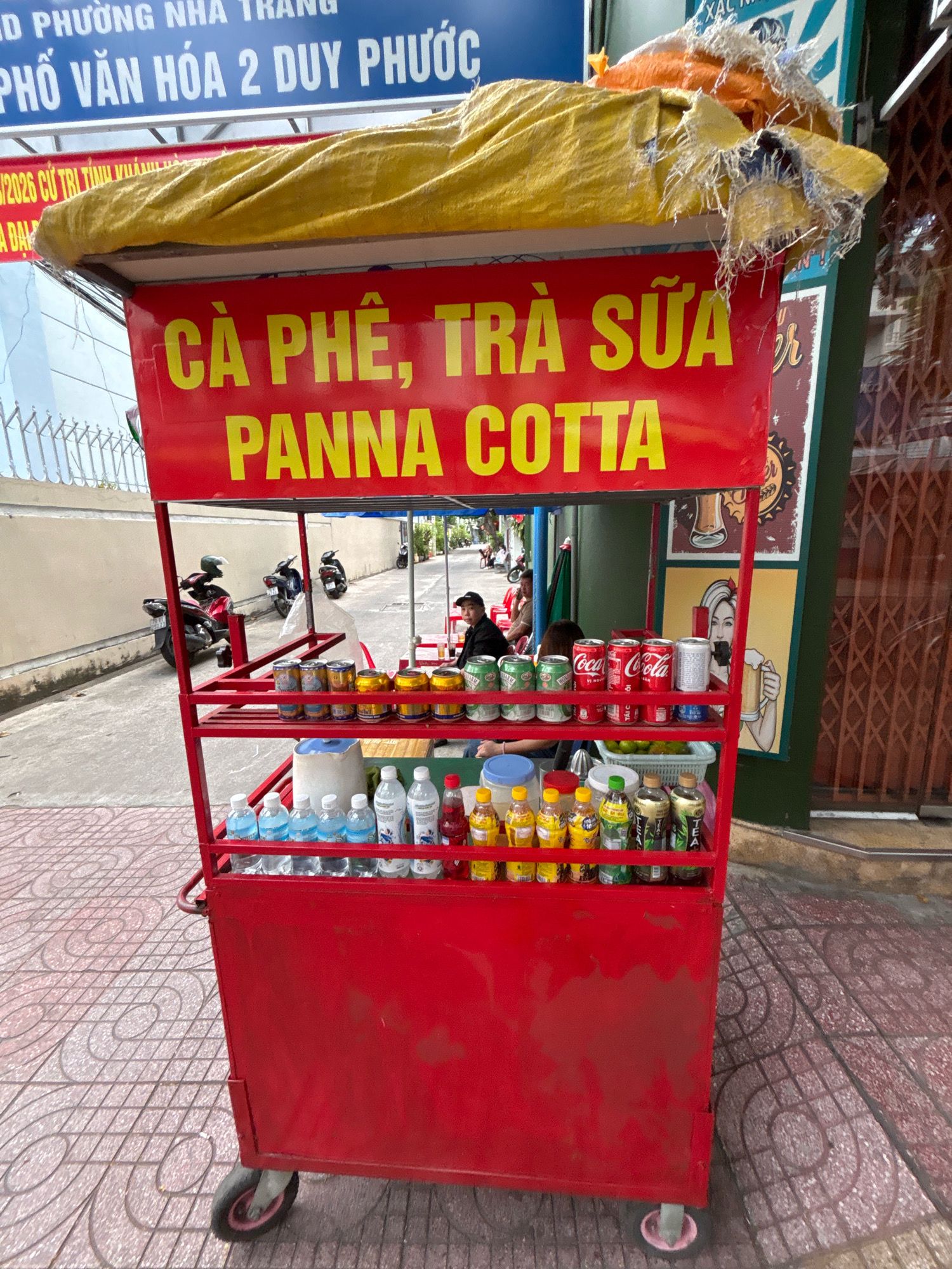 The image shows a red food cart on a sidewalk in Nha Trang, Vietnam. The cart has a yellow tarp on top and a red sign with yellow lettering that reads "CÀ PHÊ, TRÀ SỮA PANNA COTTA". The cart is stocked with drinks, including bottles of water, cans of soda, and bottles of tea. A person is seated behind the cart. In the background, there are motorcycles parked on the street and buildings. The sidewalk is paved with patterned tiles. The scene appears to be in an urban area.
