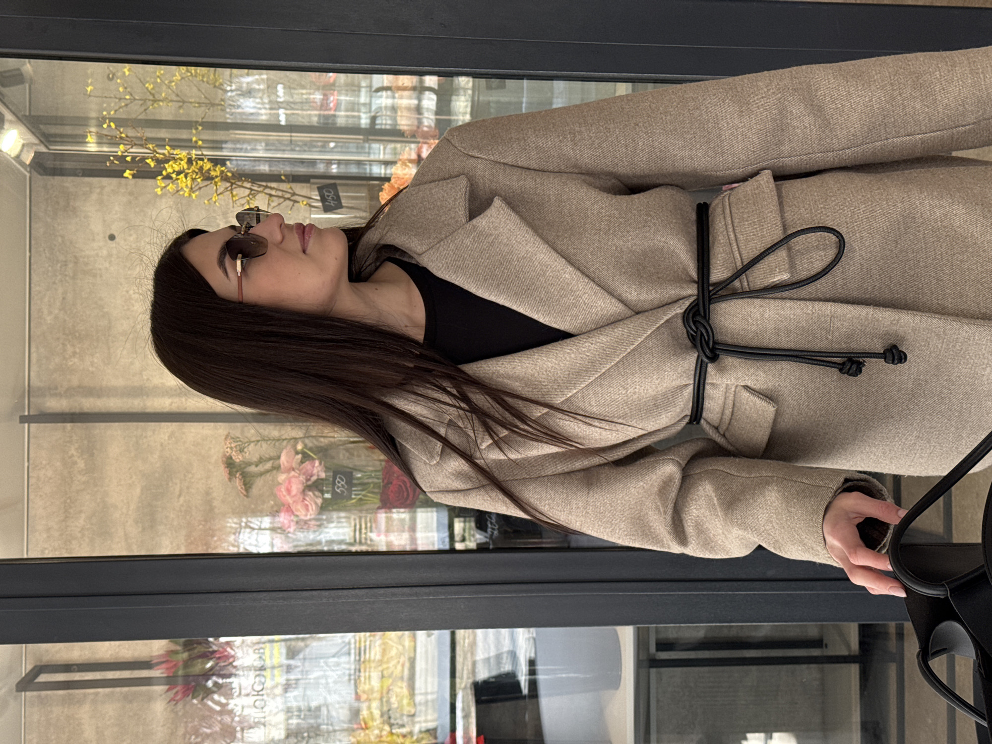 A woman with long dark hair and sunglasses is wearing a light brown coat with a black drawstring tie. She is standing in front of a display window filled with flowers, possibly in a flower shop. The window reflection reveals a city scene. Based on the Cyrillic characters visible in the reflections, the city is Chaikovsky, Russia. Other notable elements include a black top under the coat and a black handbag held in her right hand.