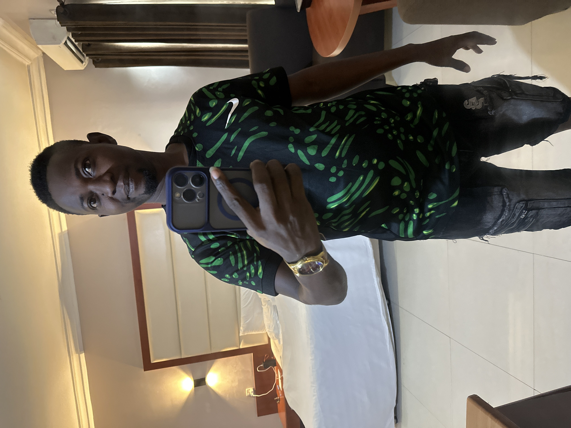 A man is standing in a hotel room, taking a selfie. He is wearing a black t-shirt with a green abstract pattern and a gold watch on his left wrist. He is also wearing distressed black denim shorts. The room has a large bed with white bedding, a headboard, two wall-mounted lights, a bedside table with some items on it, and a large window with brown curtains. An air conditioning unit is mounted high on the wall. A round wooden table and a sofa are partially visible in the background. The floor is tiled. The man is looking directly at the camera. The image appears to have been taken in the city of Abuja, Nigeria.