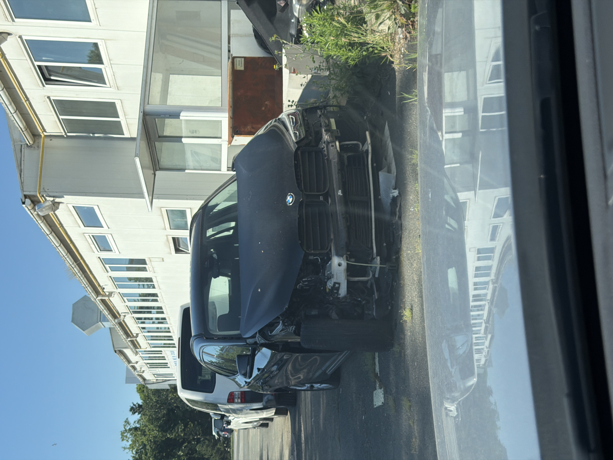 A black, damaged BMW SUV is parked in a lot next to a long, white modular building. The front of the BMW is heavily damaged, with the bumper and grille missing and the hood bent. A white van is parked behind the BMW, and other vehicles are visible in the background. The modular building has numerous windows and a yellow pipe running along its exterior. The lot appears to be paved, with some grass and debris scattered around. The scene is illuminated by bright sunlight. The building is located in Burgas, Bulgaria.