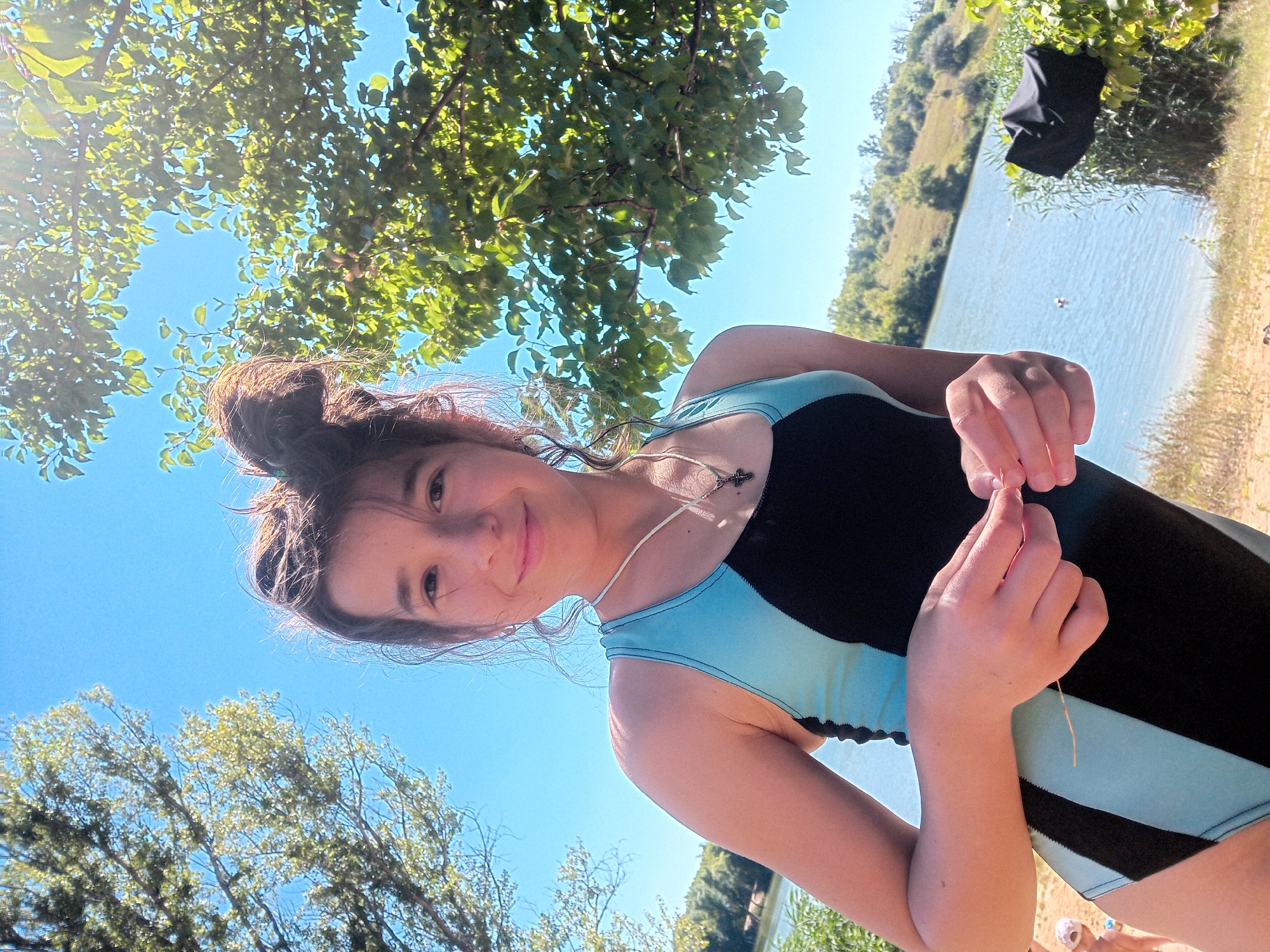 A young person is taking a selfie outdoors with a lake and hilly landscape in the background. The person is wearing a black and teal swimsuit and a necklace with a cross pendant. Their hair is pulled up in a bun, and they are smiling at the camera. The sun is shining brightly, creating lens flare in the upper right corner of the image. Green trees and foliage are visible on either side of the frame, with a clear blue sky above. The ground in the foreground appears to be sandy or dry earth. In the distance, there are a few small figures in the water, possibly swimming, and a black bag is visible amongst the bushes on the right. The overall scene suggests a warm, sunny day at a natural body of water. The location is in or near the city of Chuhuiv, Ukraine.