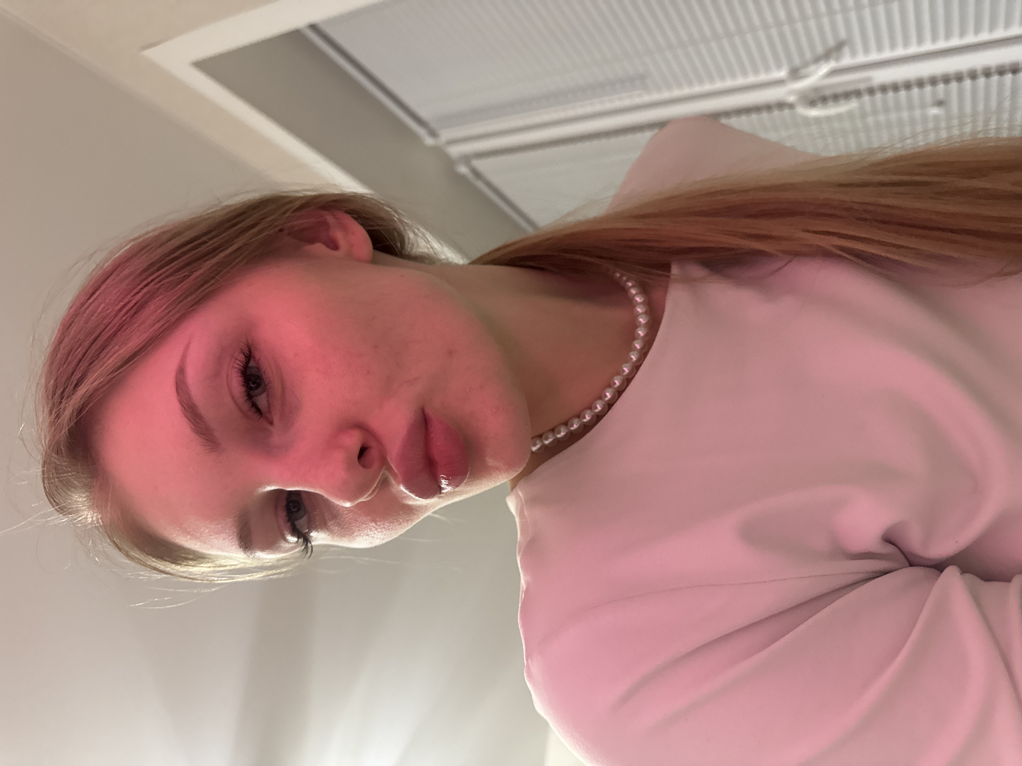 A close-up, rotated portrait shot captures a young woman looking downwards at the camera from a low angle. She has long, light brown hair and is wearing a light-colored top, possibly white or light pink, accessorized with a pearl necklace. Her face is prominently featured, with her eyes open and eyelashes visible. Her lips are slightly parted and appear glossy.
A strong pink or red light source illuminates the left side of her face and hair, creating a distinct hue, while the right side appears more naturally lit. The background is softly out of focus, revealing an indoor setting with a white ceiling structure, possibly a ventilation grid or panel. No specific activity beyond posing for the photo is depicted. The artificial, colored lighting suggests an evening or controlled indoor environment. The image is devoid of any visible text. The overall impression is one of a styled, intimate portrait, emphasizing the subject's features and the dramatic lighting. The location context for this media file is Perm, Russia.