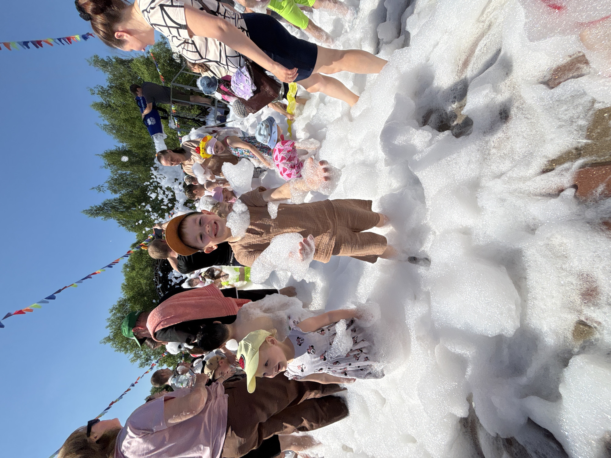 A group of people, mostly children, are enjoying a foam party in an outdoor setting under a clear blue sky. The ground is covered in a thick layer of white foam, with some areas showing glimpses of brickwork underneath. The children are dressed in casual summer clothing, and some are wearing hats. One boy in the foreground, wearing a brown outfit and a cap, is smiling and holding up foam in his hands. To his left, a young girl in a patterned dress and a yellow cap is looking to the side and also surrounded by foam. In the background, adults are visible, some watching the children and others possibly participating. There are colorful flags strung across the area, suggesting a festive event. A man in the background is operating a foam machine, creating more foam. The overall scene depicts a fun, celebratory atmosphere. The location is identified as a public space in the city of Заволжье, Russia.