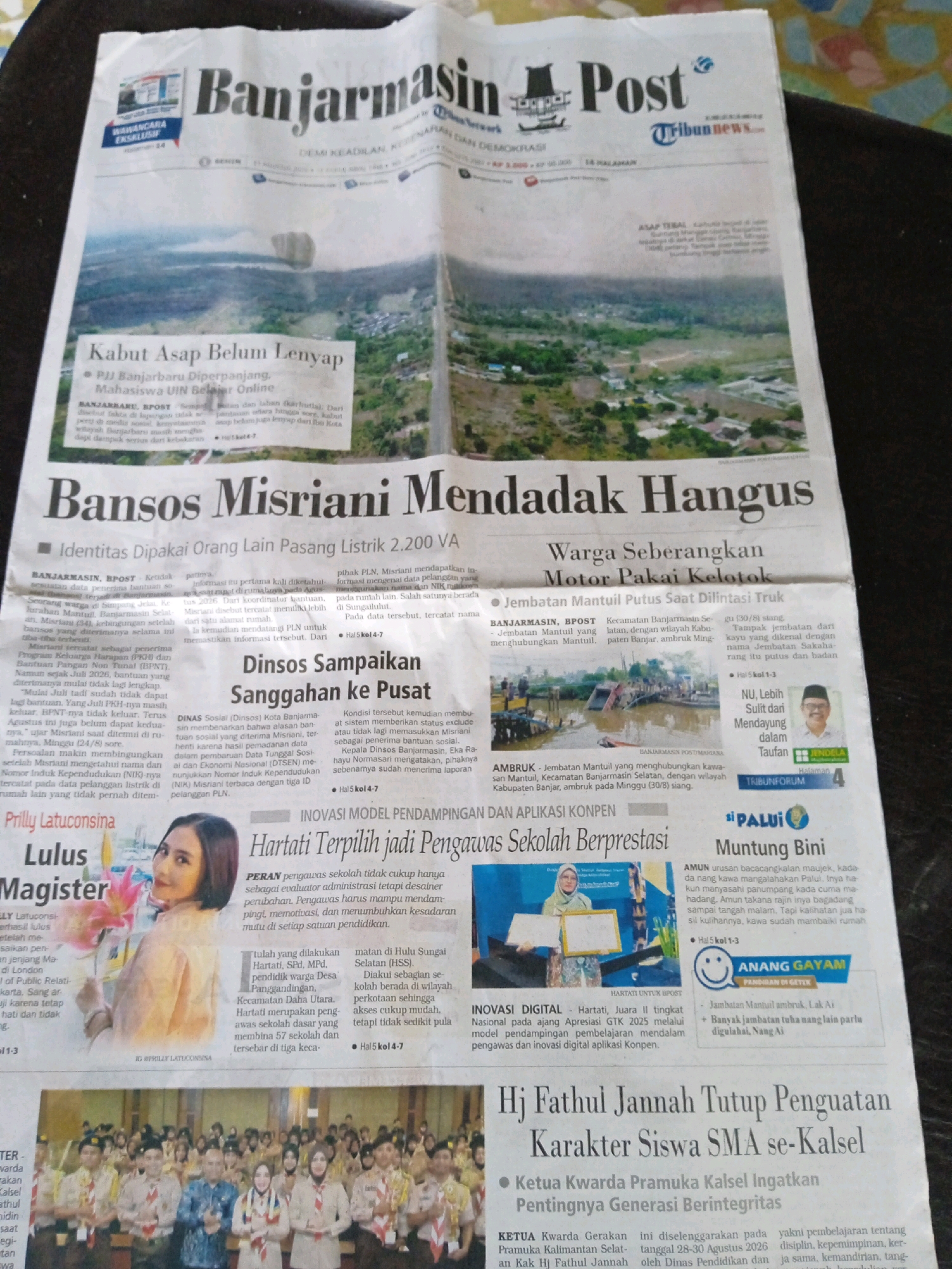 The image is a close-up, top-down view of a newspaper, the "Banjarmasin Post," dated Friday, August 28, 2020. The newspaper is spread out, with the masthead prominently displayed at the top. Several articles are visible, covering local news and events in Banjarmasin, Indonesia.
The main headline on the left side of the paper reads "Kabut Asap Belum Lenyap" (Haze Has Not Disappeared Yet), with a sub-headline about PJJ Banjarbaru Diperpanjang, Mahasiswa UIN Belajar Online (PJJ Banjarbaru Extended, UIN Students Study Online). Further down, a large headline declares "Bansos Misriani Mendadak Hangus" (Misriani's Social Aid Suddenly Burns Up), with a sub-headline "Identitas Dipakai Orang Lain Pasang Listrik 2.200 VA" (Identity Used by Others to Install 2,200 VA Electricity). Another article discusses "Dinsos Sangkaikan ke Pusat" (Social Services Suspect to the Center).
On the right side, a headline states "Warga Seberangkan Motor Pakai Kelotok" (Residents Cross the River Using a Motorboat), with a sub-headline "Jembatan Mantuil Putus Saat Dilintasi Truk" (Mantuil Bridge Collapsed When Crossed by a Truck). Below this, an article titled "NU, Lebih Sulit Mendayung dalam Taufan" (NU, Harder to Row in a Storm) is accompanied by a photo of a man. Further down, a photo of a woman with flowers is captioned "Prilly LatucConsina Lulus Magister" (Prilly LatucConsina Graduates with a Master's Degree).
The bottom section of the newspaper features a prominent headline "Hj Fathul Jannah Tutup Penguatan Karakter Siswa SMA se-Kalsel" (Hj Fathul Jannah Closes Character Strengthening for High School Students in South Kalimantan). This is accompanied by a photograph of a group of students and adults in uniform, possibly at an event.
The overall impression is of a local newspaper reporting on a variety of community issues, from environmental concerns like haze to social services, infrastructure, and educational achievements. The use of bold headlines and accompanying photographs aims to attract reader attention to the featured stories.