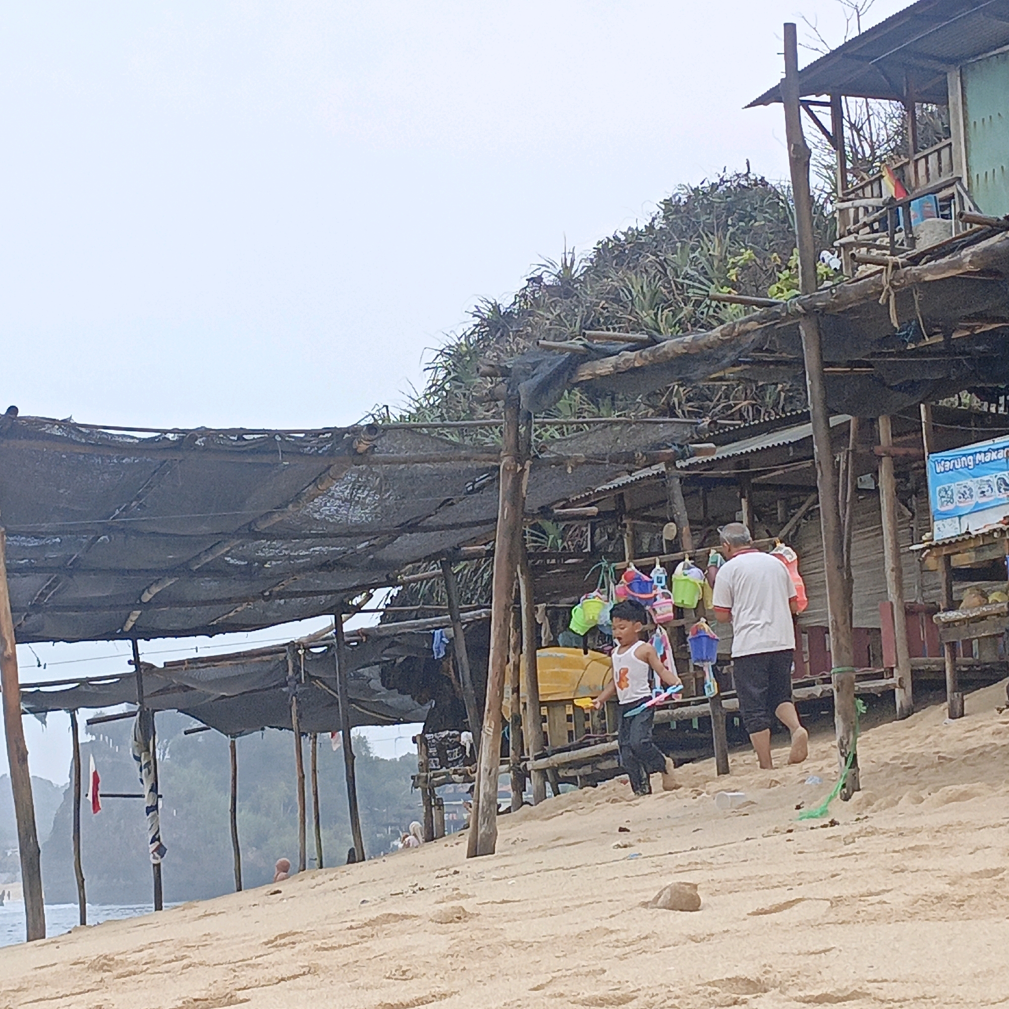 The image depicts a beachfront scene in Tepus, Indonesia, featuring a rustic, open-air establishment. The structure, supported by wooden poles, is partially covered by a dark tarp and a corrugated metal roof. Displayed on hooks are various colorful buckets and toys. A young boy in a white tank top and dark pants is walking away from the viewer, holding a pink and blue toy. Behind him, an older man in a white shirt and dark pants is also walking away. The ground is sandy, and the ocean is visible in the background. The overall impression is one of a casual, seaside vendor or eatery.