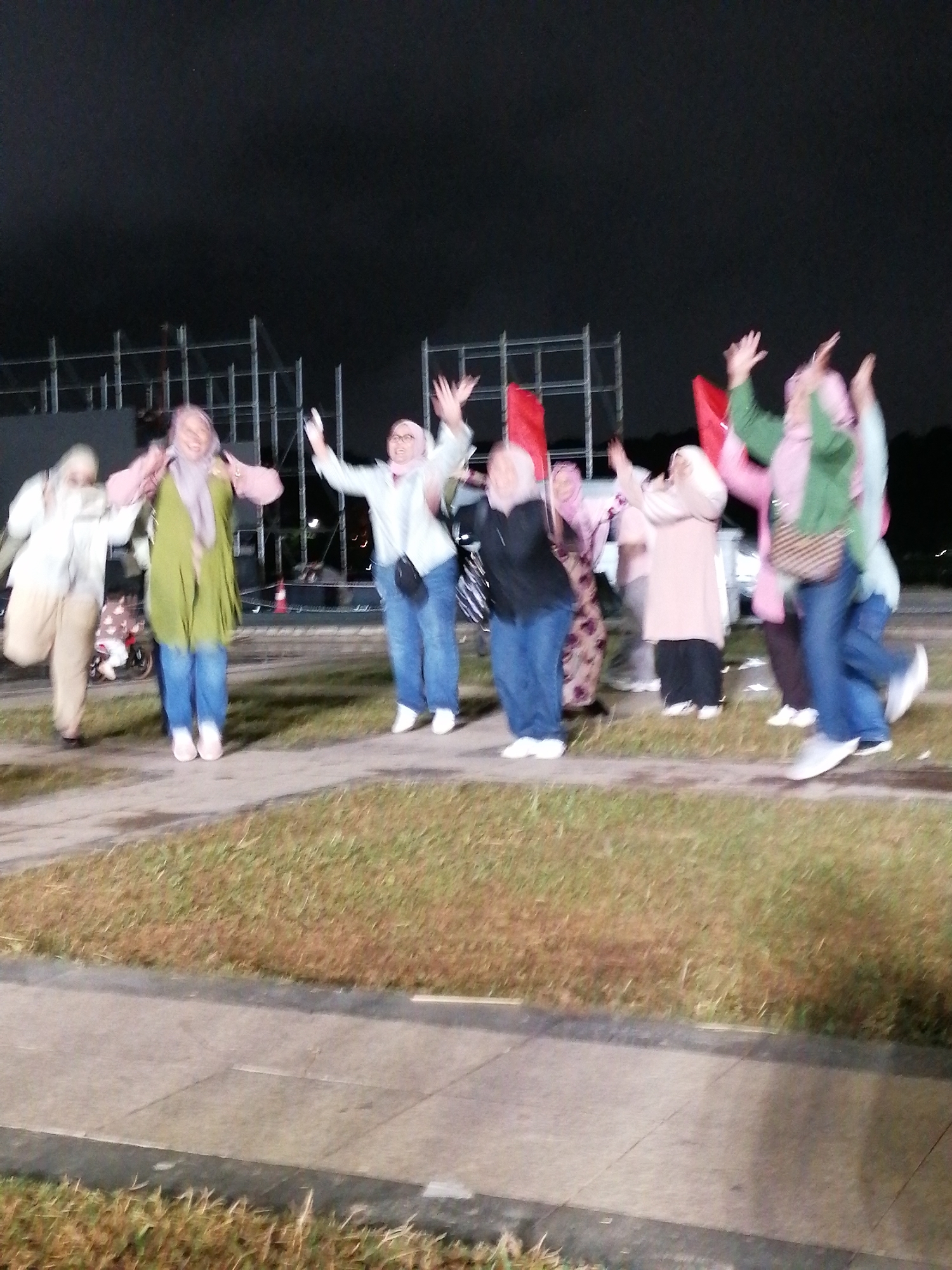 The image depicts a group of approximately ten individuals, mostly women, outdoors at night. They appear to be in a celebratory mood, with several individuals jumping and raising their arms. Some are holding red flags or fabric. The individuals are wearing casual attire, including jeans, long shirts, and headscarves (hijabs). The background shows dark, indistinct structures, possibly scaffolding or buildings, and some low-lying vegetation and pavement in the foreground. The lighting suggests artificial illumination from the event. The presence of red flags may indicate a nationalistic or celebratory event. The image does not contain any specific visual cues that can definitively place the scene in Da Lat, Vietnam.
