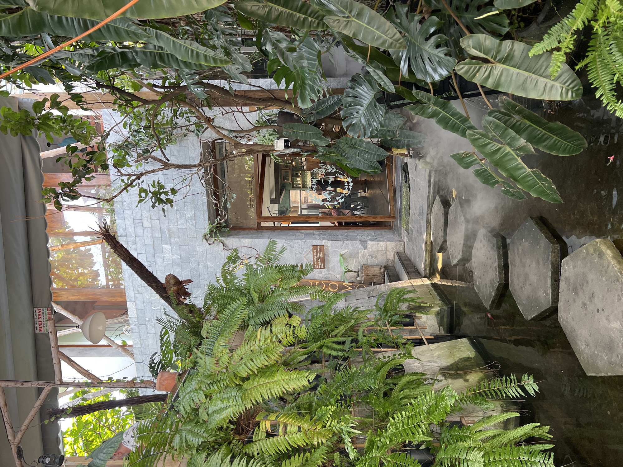 The photograph shows a building entrance located in Nha Trang, Vietnam. The building is constructed from gray brick. The doorway is framed in wood. A pond containing stepping stones is situated in front of the entrance. The scene is heavily populated with greenery, including ferns and large-leafed plants. Vapor is present near the pond. People are visible inside the building, behind the glass door. A sign is mounted on the wall beside the entrance. Exterior lighting fixtures are also visible.