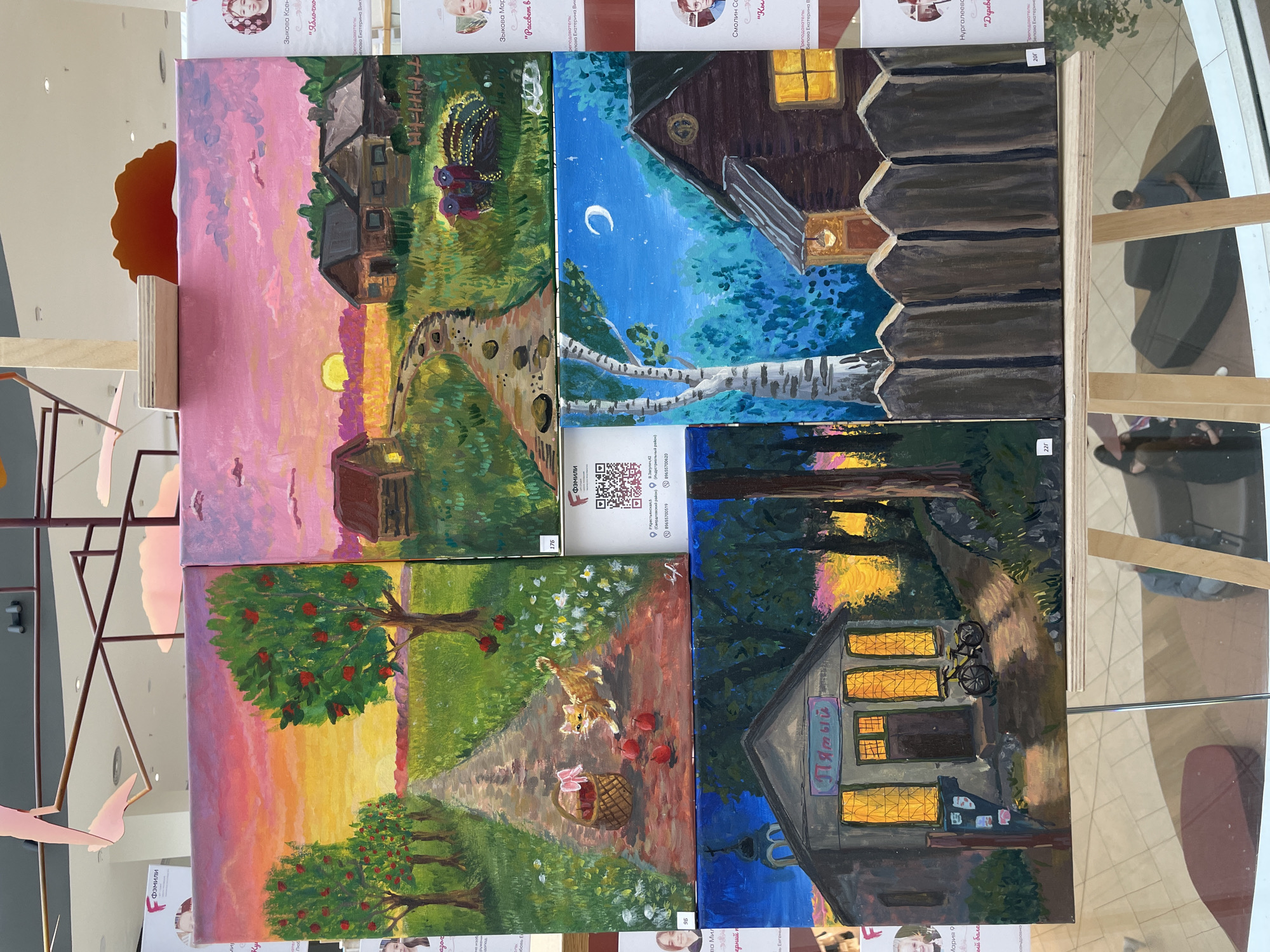 This image displays a collection of four acrylic paintings, arranged in a 2x2 grid, presented at an exhibition in Perm, Russia. The paintings depict idyllic rural scenes, likely rendered by children or young artists, as suggested by the style and some of the visible text, which includes names like "Зыкова Ксен", "Зыкова Мар", "Смолин Се", and "Нургалеевс", along with titles such as "Яблочно", "Рассвет в", and "Дерев".
The top-left painting captures a rustic village scene at sunset or sunrise, with a warm, pink-hued sky illuminating a wooden house and surrounding greenery. Two turkeys are prominent in the foreground, adding a touch of farm life. The path leading towards the house suggests a journey or arrival.
The top-right painting shifts to a night scene, showcasing a wooden house under a deep blue, star-filled sky with a crescent moon. A lone window emits a warm yellow light, hinting at a cozy interior. The natural setting is characterized by lush, dark foliage and the distinctive white bark of birch trees.
The bottom-left painting depicts a vibrant orchard with a red-apple-laden tree and a path winding through a grassy field dotted with white flowers. A small, fluffy cat is playfully chasing apples, with a charming straw basket and a pink bow placed nearby. This scene evokes a sense of innocence and playful activity.
The bottom-right painting presents a small building, possibly a shop or a house, illuminated by warm light from its windows. A bicycle is parked outside, suggesting recent activity. The building features a sign with Cyrillic lettering that reads "Пятый", which translates to "Fifth", likely indicating its name or number. The overall mood across all paintings is one of tranquility, nostalgia, and a gentle appreciation for rural life. The reflections on the glass surface indicate that these paintings are displayed behind a protective barrier.