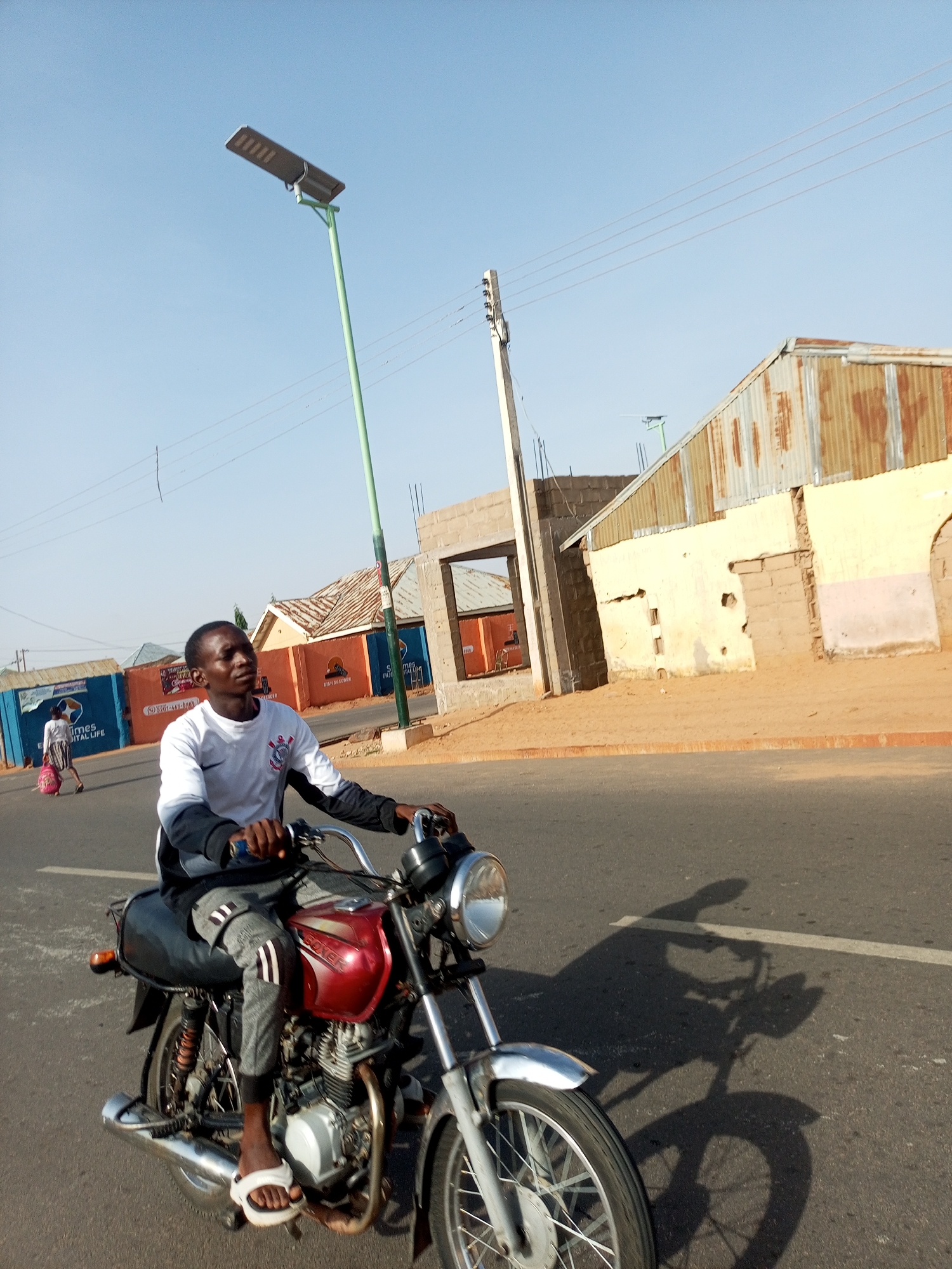 The image depicts a man riding a red and silver motorcycle on an asphalt road in what appears to be the city of Gombe, Nigeria. The man is wearing a white t-shirt with long black sleeves, grey pants with white stripes, and white sandals. He is looking to his left. The motorcycle has a black seat and chrome handlebars.
In the background, there are buildings of varying construction styles. One building has a corrugated metal roof, and another is made of concrete blocks. A green streetlight stands tall. There is another person walking in the background with an indiscernible pink object. Power lines are visible in the sky. The environment suggests a sunny day.