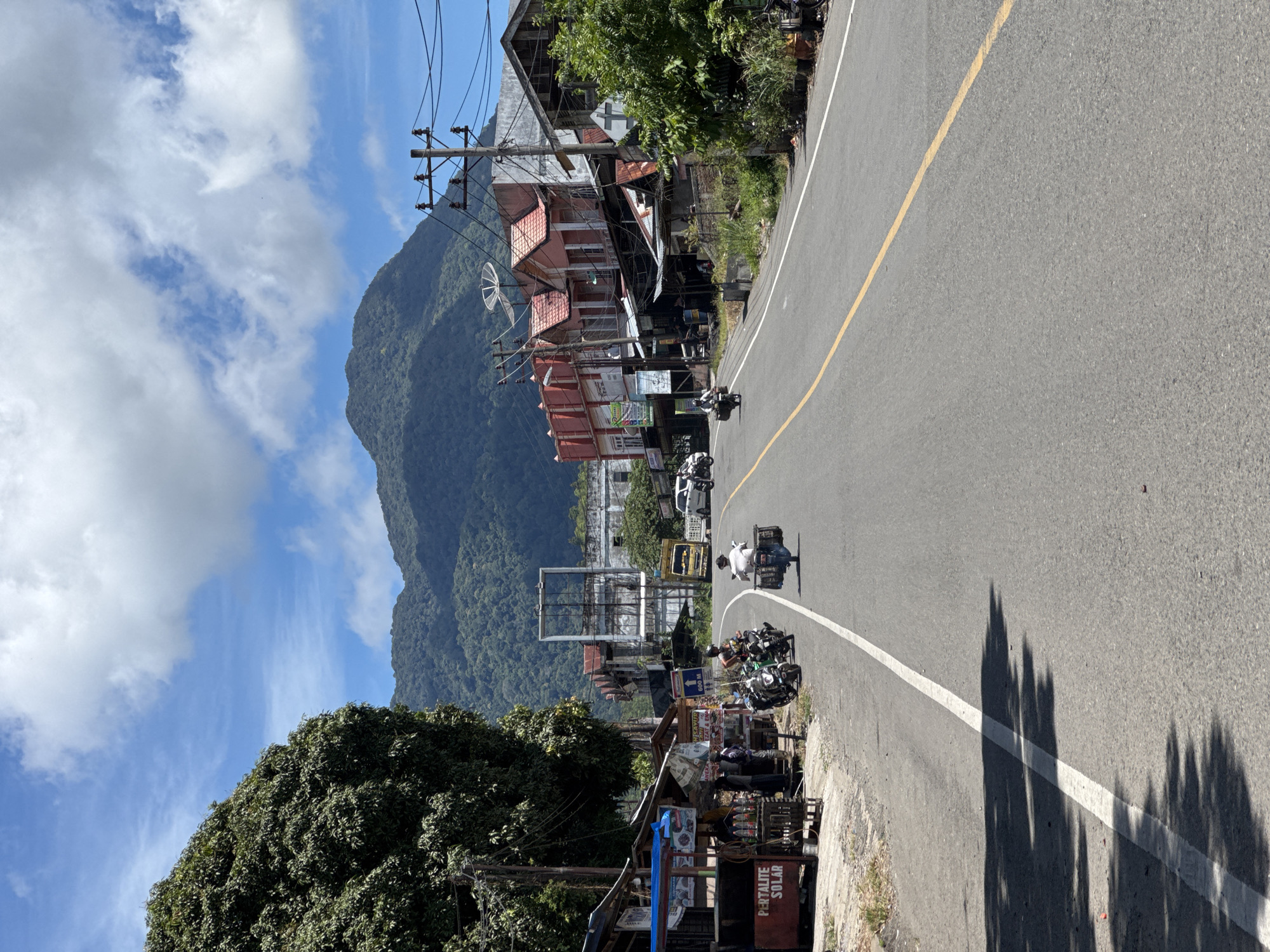 This is a medium-angle, eye-level shot of a road in Saree, Aceh, Indonesia, on a bright, sunny day with scattered clouds. The road curves slightly to the right and slopes gently uphill. On the left side of the road, there are several small shops and stalls, some with blue awnings, and a large tree with dense foliage. A sign on one of the stalls reads "PERTALITE SOLAR". Several motorcycles are parked near these shops.
Further down the road, on the right, are more buildings, some with red-tiled roofs and others appearing more modern. There are utility poles with numerous wires running overhead. In the background, lush green mountains rise against the blue sky.
There are several people and vehicles visible. A person on a motorcycle with a sidecar is traveling away from the camera in the right lane. Two people on motorcycles are stopped or moving slowly near the shops on the left. Another person is seen walking on the sidewalk on the left. In the distance, a yellow truck and a white car are on the road. The overall scene depicts a lively street in a mountainous region.