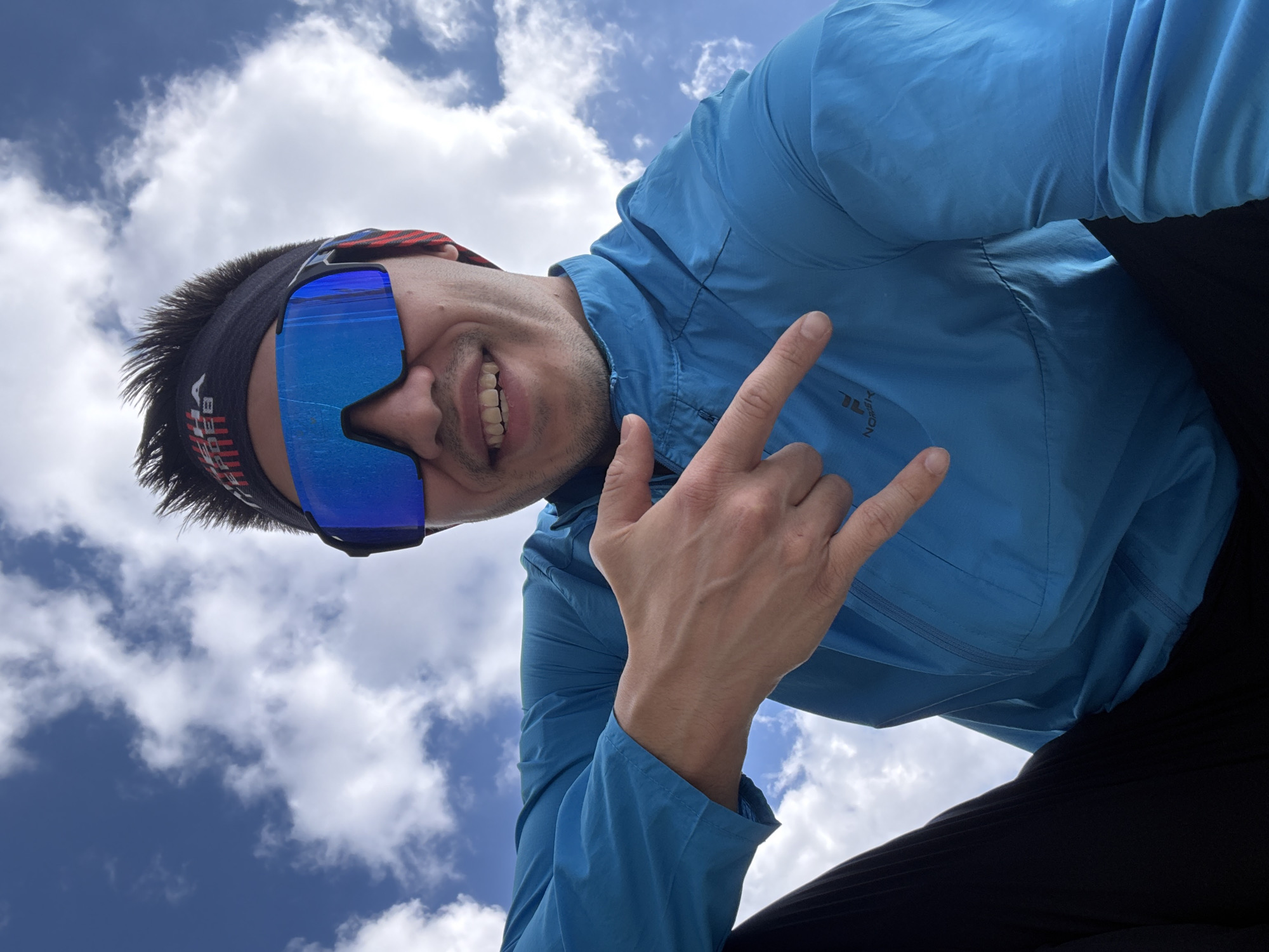 A low-angle shot of a smiling man with a blue jacket, black headband and blue sunglasses set against a backdrop of a blue sky dotted with white clouds. The man is giving a "rock on" hand gesture. The man is likely located outdoors but precise location information like the city of Kazan, Russia is not discernible from the image alone.