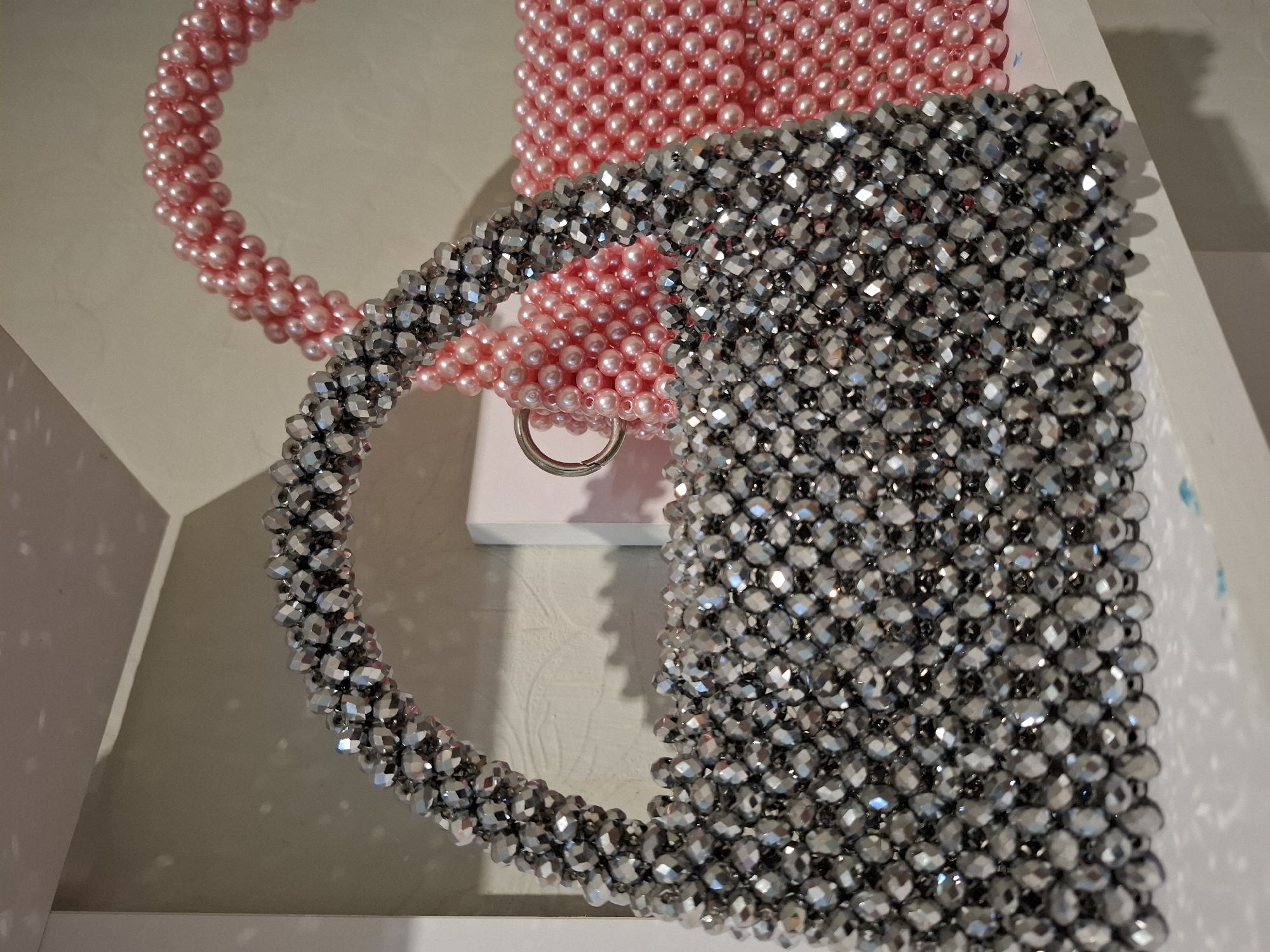 The image displays two beaded handbags on a white shelf. The handbag in the foreground is silver and heavily embellished with faceted beads. The handbag behind it is pink and appears to be made of smaller, smoother beads. A silver ring is attached to the pink handbag. The background is a light-colored wall with subtle shadows. The scene suggests a retail or display environment. The location of Korenovsk, Russia, is not visually represented in the image.