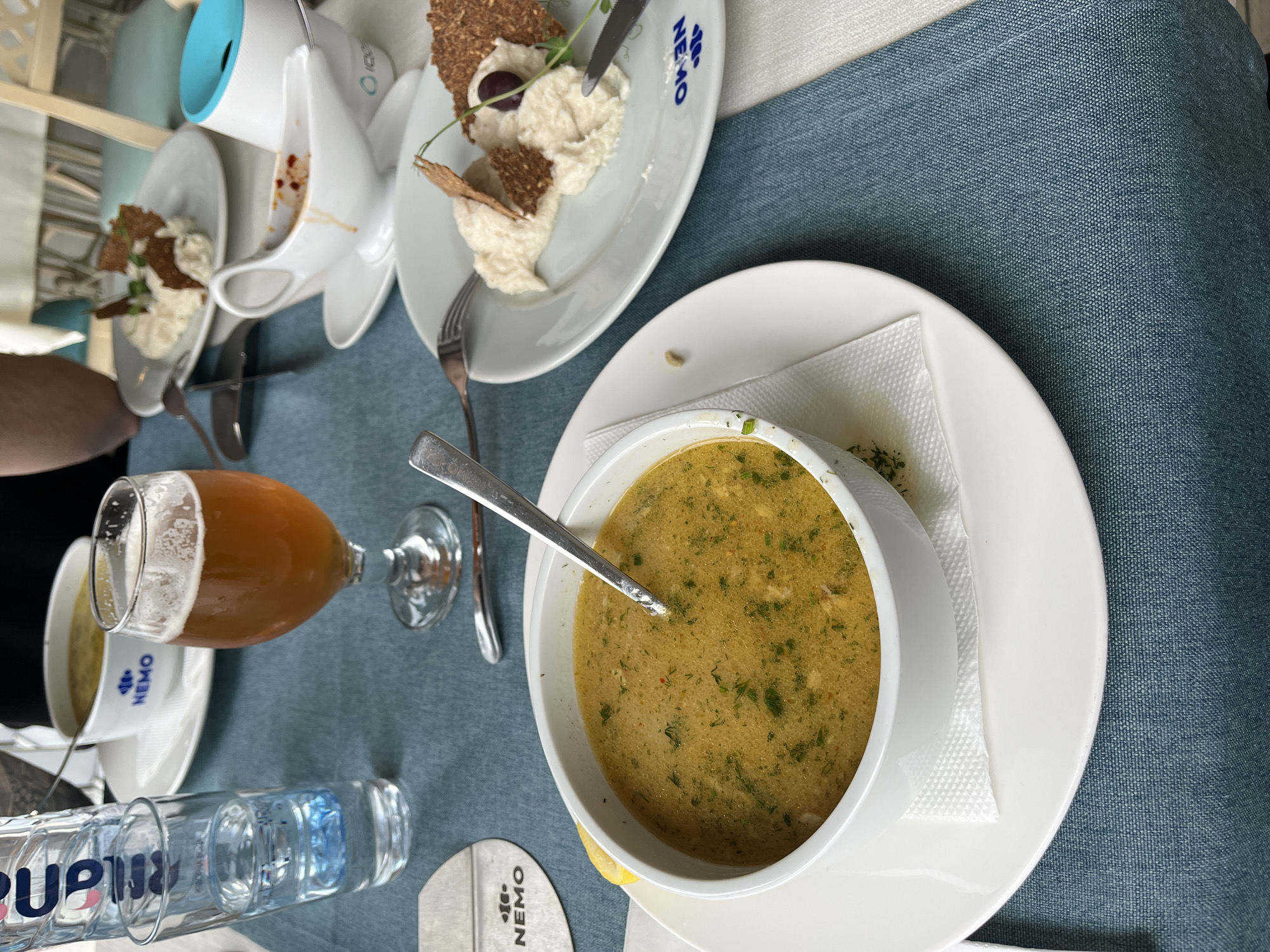 The image shows a table setting at a restaurant, presumably in Varna, Bulgaria given the "NEMO" branded items. On the table are dishes with what appear to be appetizers or starters. A bowl of soup is also present, along with a glass of beer and a bottle of water. The table is covered with a blue linen cloth. Part of a person's arm and shoulder can be seen on the left side of the image.