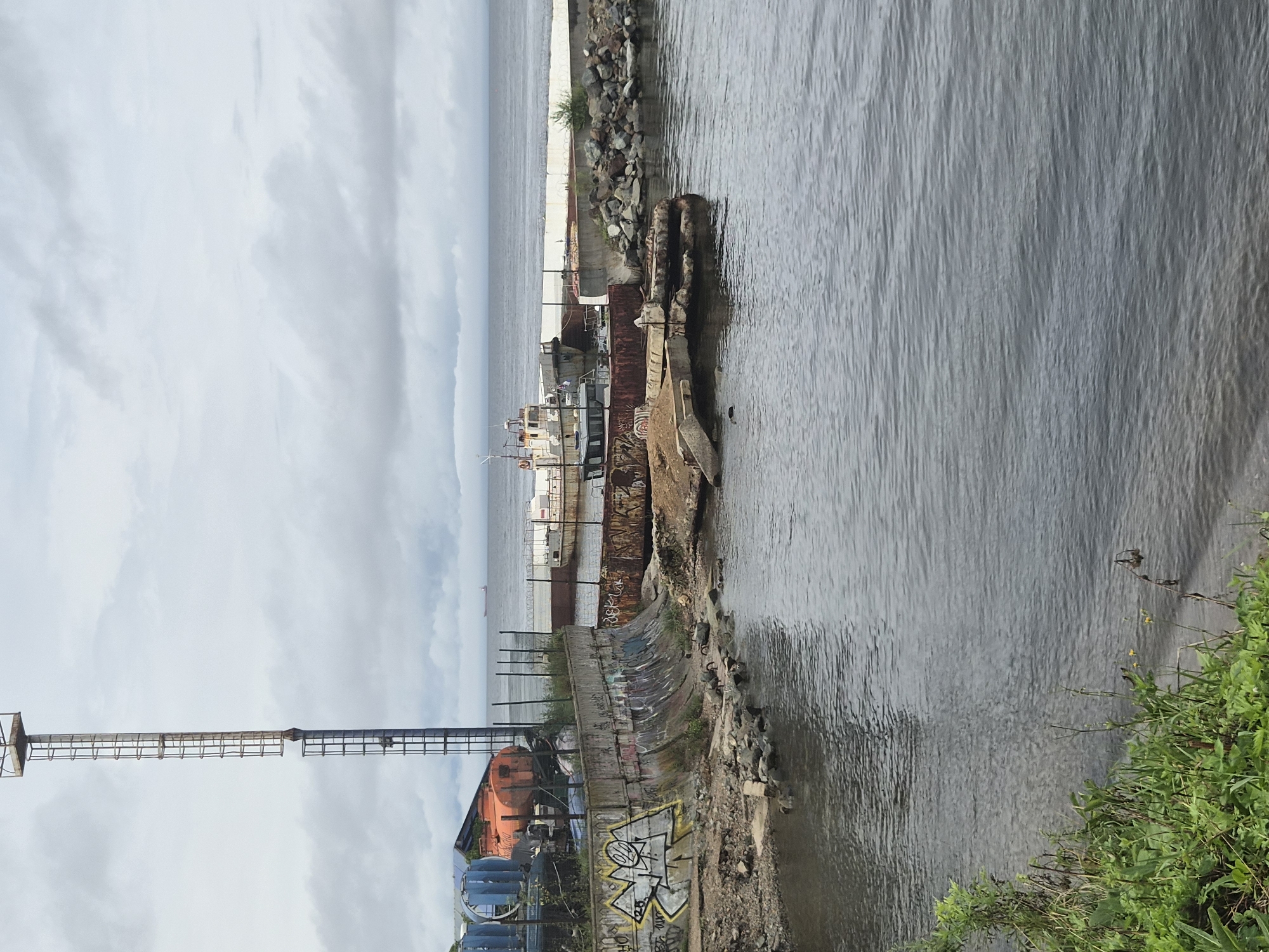 A cloudy day is depicted at a dock in Vladivostok, Russia. In the foreground, the calm, grey water laps at a grassy embankment on the left. A concrete retaining wall, covered in graffiti, runs along the midground, separating the water from a derelict industrial area. Various rusted metal structures, including a large orange tank and pipes, are visible in the background, along with parts of a boat. A tall, metal tower, possibly a crane or observation post, stands prominently on the left. In the distance, the sea stretches to the horizon under a grey sky, with a few boats visible. The scene suggests an abandoned or disused part of the harbor.