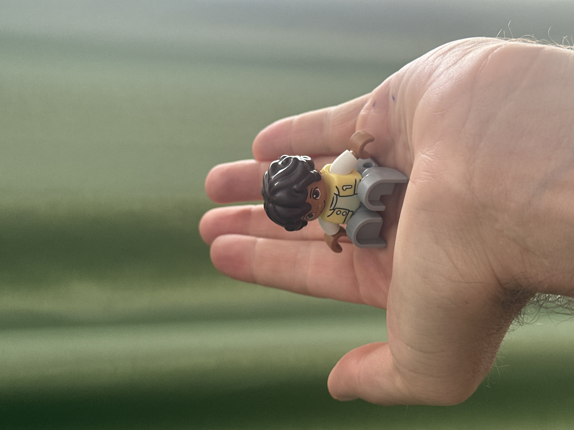 A Lego minifigure of a person with brown hair, a yellow vest over a white shirt, and gray pants is seated in the palm of a person's hand. The hand is open, with the fingers curved upwards, supporting the minifigure. The background is a green, vertical textured surface, possibly a curtain or drapery, with soft lighting. The image is shot from a slightly elevated perspective, focusing on the minifigure and the hand. There are faint blue markings on the palm of the hand. There is no indication of the image being located in Fethiye, Turkey.