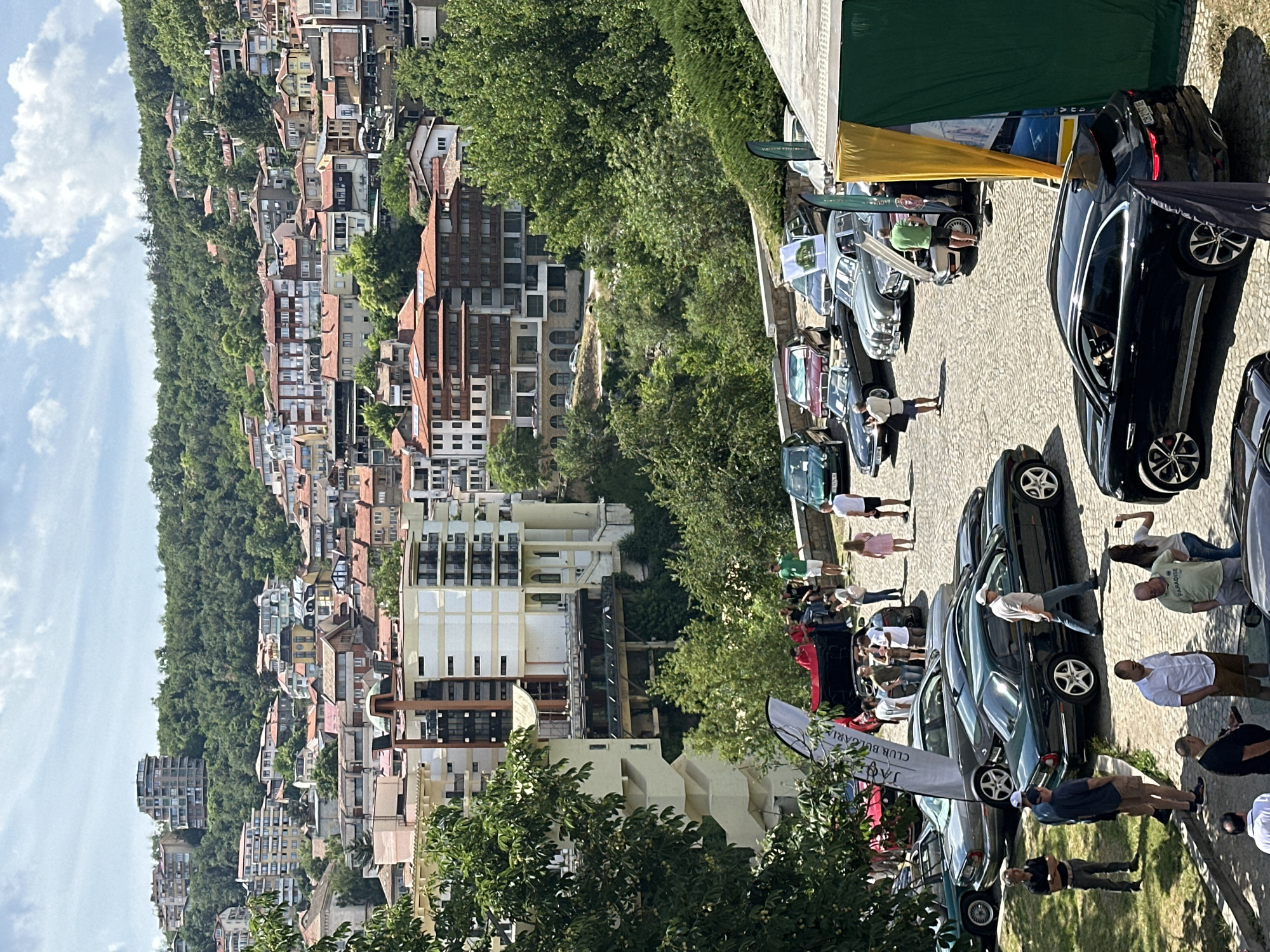 The image captures a gathering of people and classic cars in a cobblestone plaza with a backdrop of a densely populated hillside covered in buildings and lush green trees. This is the city of Veliko Tarnovo, Bulgaria, known for its historic architecture perched on steep slopes.
In the foreground, a dark-colored Jaguar F-Pace is parked, facing away from the viewer. To its left, a group of men are gathered, some conversing. Behind them, several classic cars are arranged, including a dark green Jaguar sedan, a silver Jaguar sedan, a red convertible, and a burgundy sedan. People are milling around these vehicles, some looking at the cars, others standing and talking. A tall, white banner with "JAGUAR CLUB BULGARIA" printed on it is prominently displayed near the cars.
Further back, more people are visible, including individuals dressed casually, some wearing shorts and t-shirts, suggesting a warm day. The buildings on the hillside are a mix of architectural styles, with terracotta tiled roofs and beige or white facades, characteristic of Veliko Tarnovo. The dense foliage of trees creates a vibrant green contrast against the urban landscape. The sky is partly cloudy, indicating a sunny day. The overall scene suggests an outdoor event or exhibition focused on classic cars, set within the picturesque urban environment of Veliko Tarnovo.